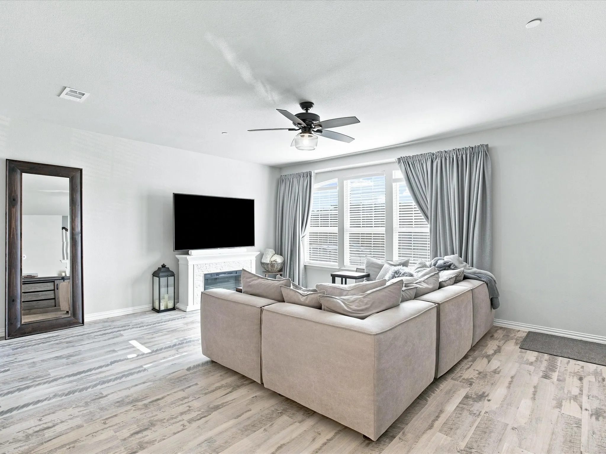 Wood-finish flooring, ceiling fan with integrated lighting, wall-mounted television, fireplace with decorative mantel, and expansive windows with blinds