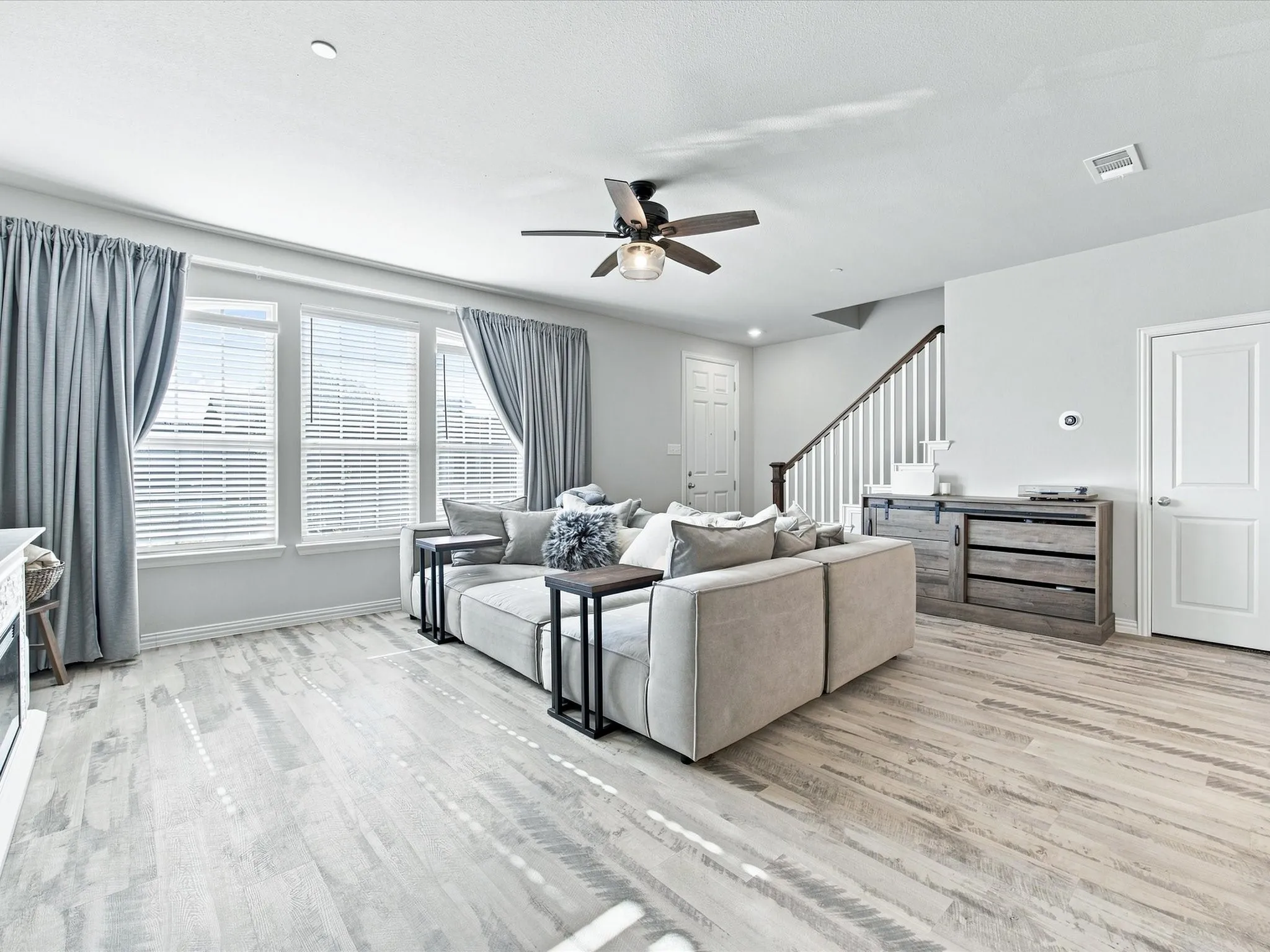 Wood-finish flooring, multiple windows with blinds, ceiling fan with integrated lighting, white interior doors, and a staircase with white balusters