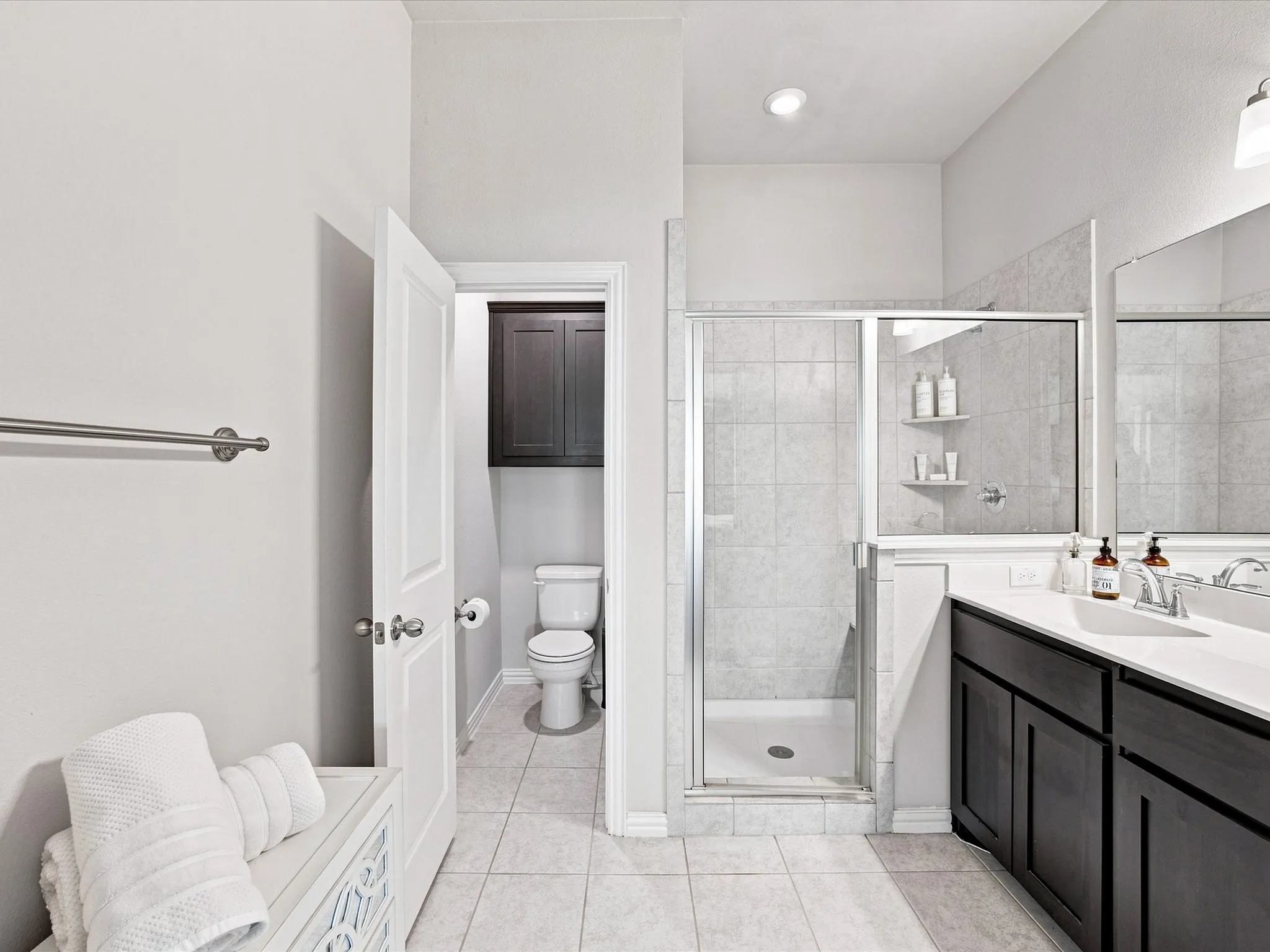 Walk-in shower with clear glass enclosure and tiled walls, dual-sink vanity with dark wood-finish cabinetry, white countertop, and integrated sink