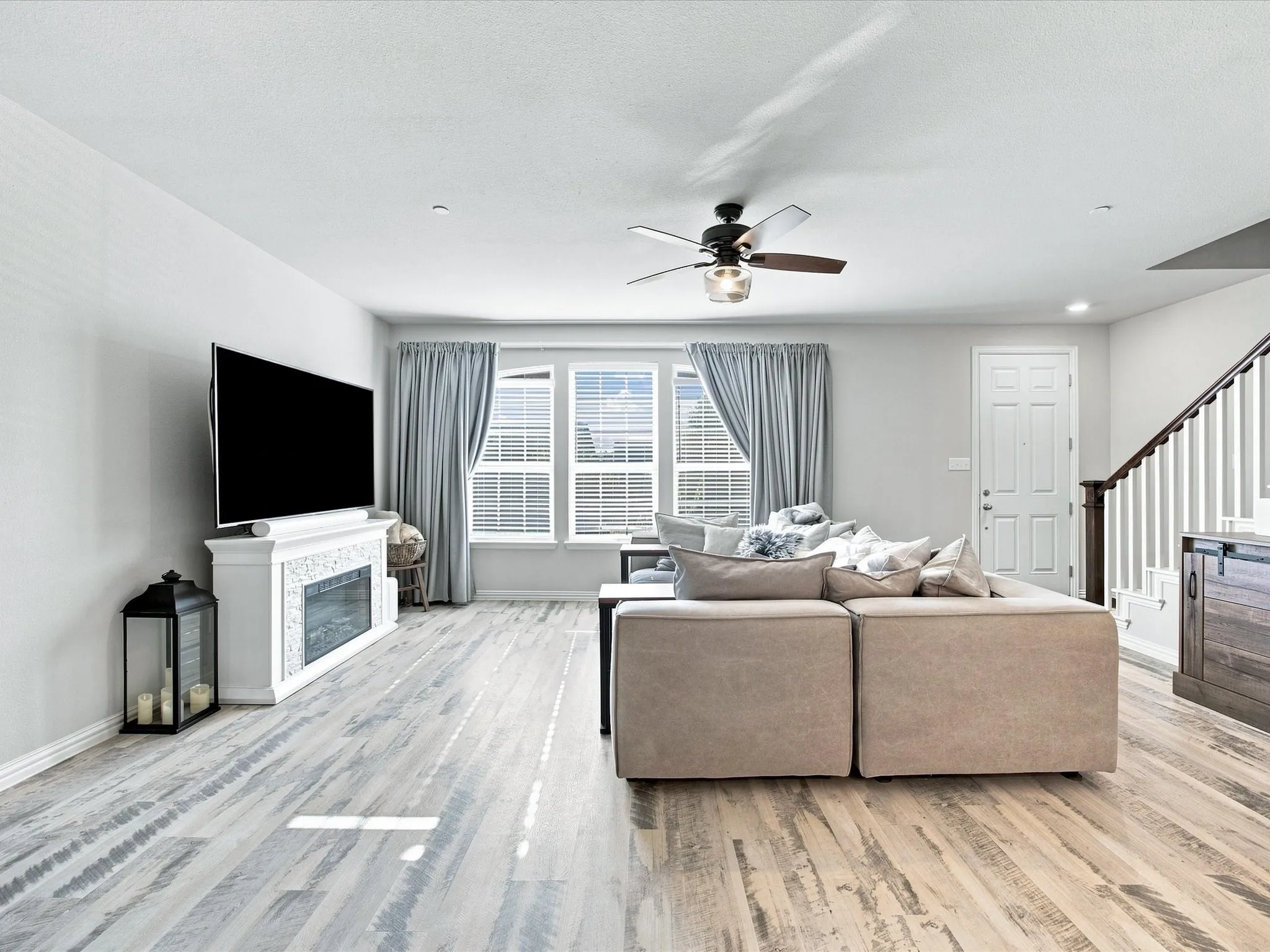Spacious living area featuring wood-finish flooring, a white mantel fireplace, multiple windows with blinds, a ceiling fan with integrated lighting, and a paneled entry door