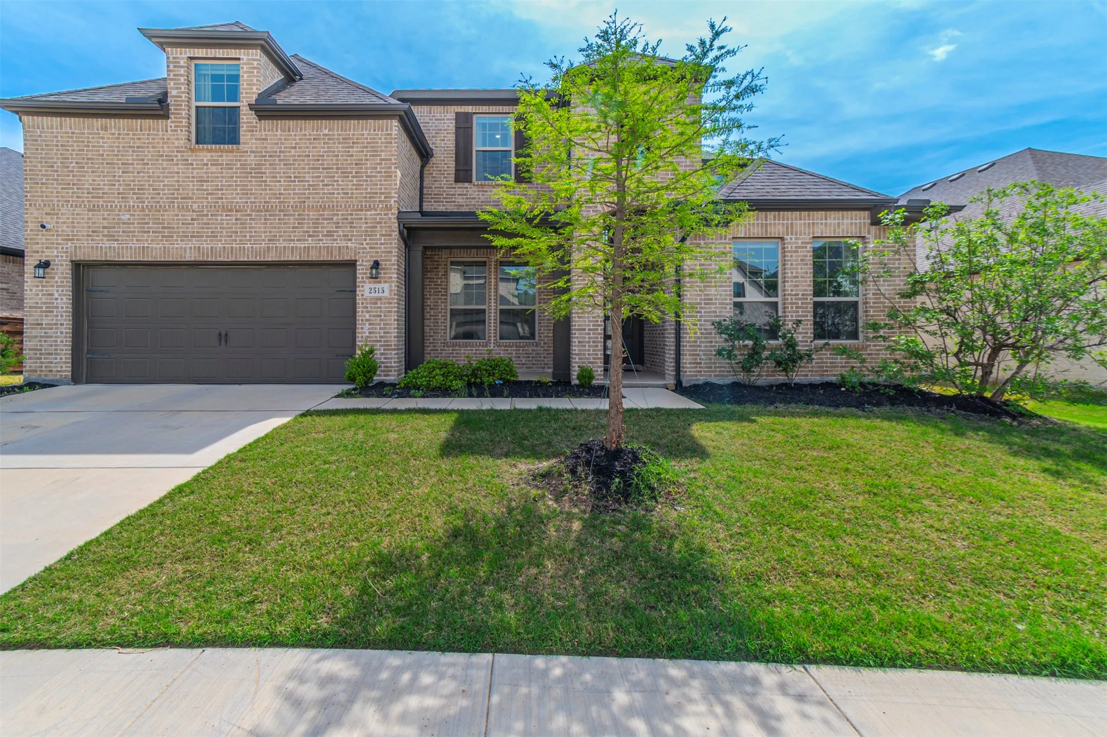 Brick facade with dark trim accents, integrated two-car garage, dormer window, and mature front landscaping