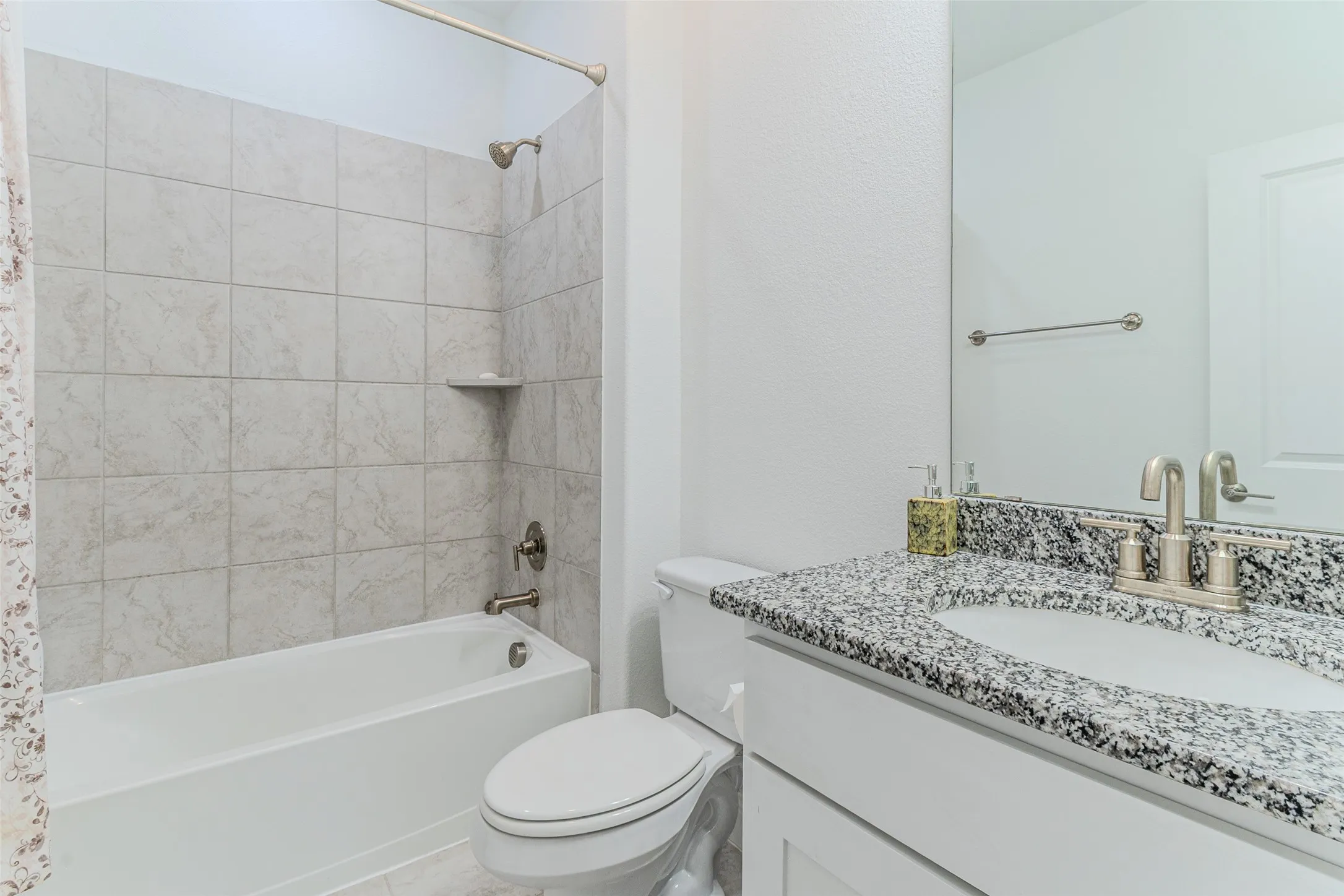 Bathtub and shower combination with light-toned tiled surround, vanity with dark speckled countertop, undermount sink, and brushed nickel faucet