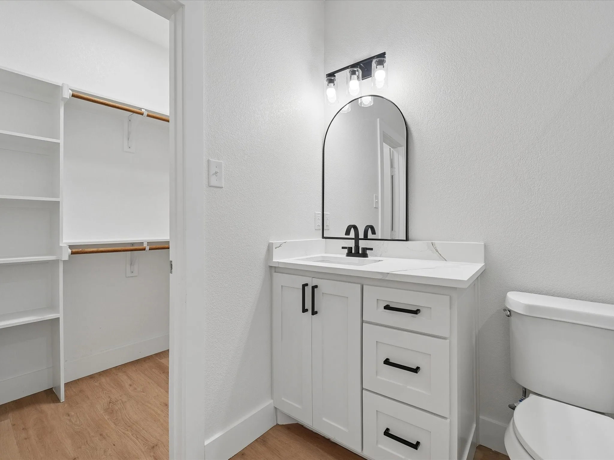 White vanity with a stone-finish countertop, black hardware, an arched black-framed mirror, and a three-light vanity fixture