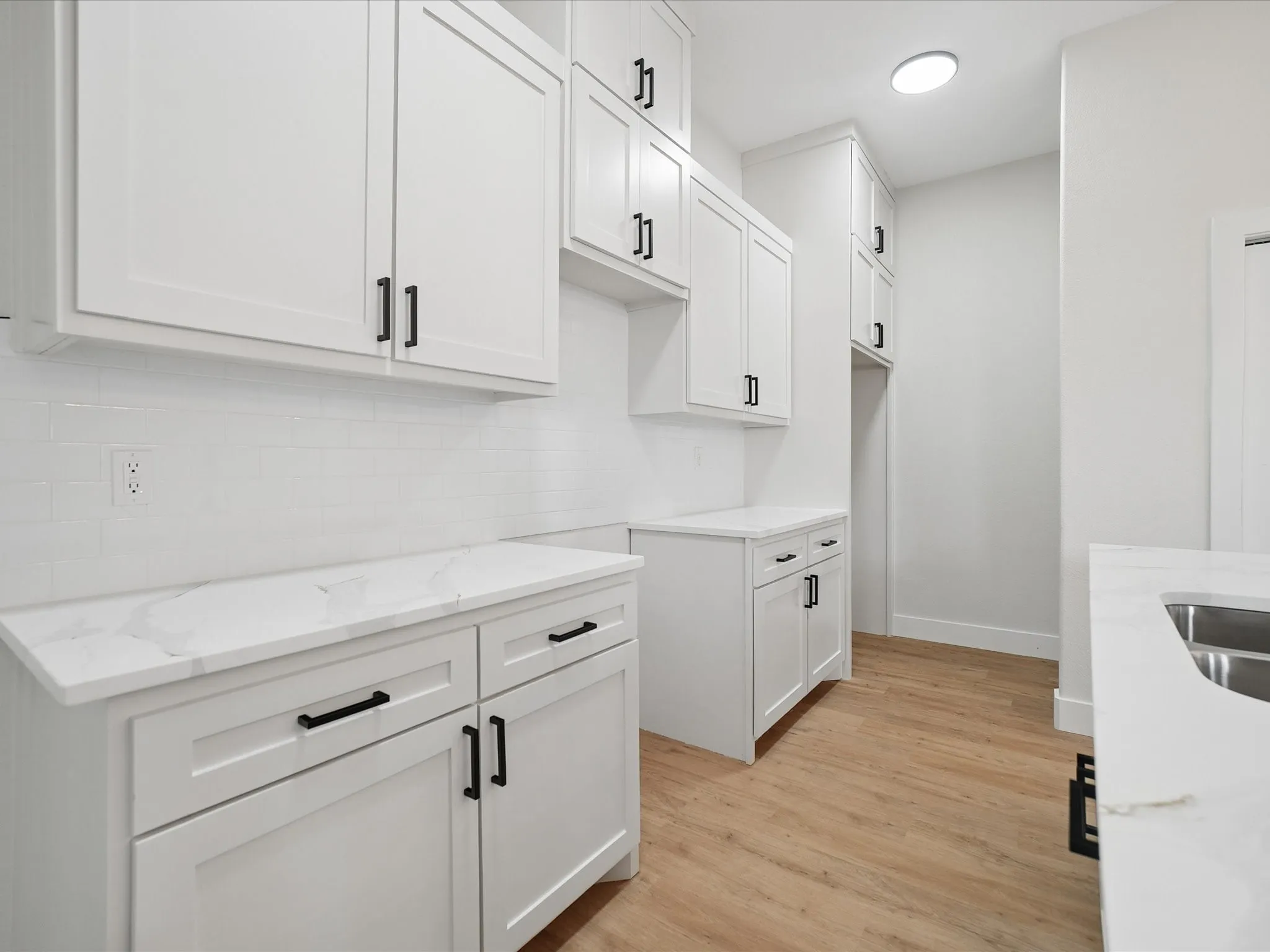 White shaker-style cabinetry with black hardware, white subway tile backsplash, white countertops with veining, wood-finish flooring, and a recessed ceiling light fixture