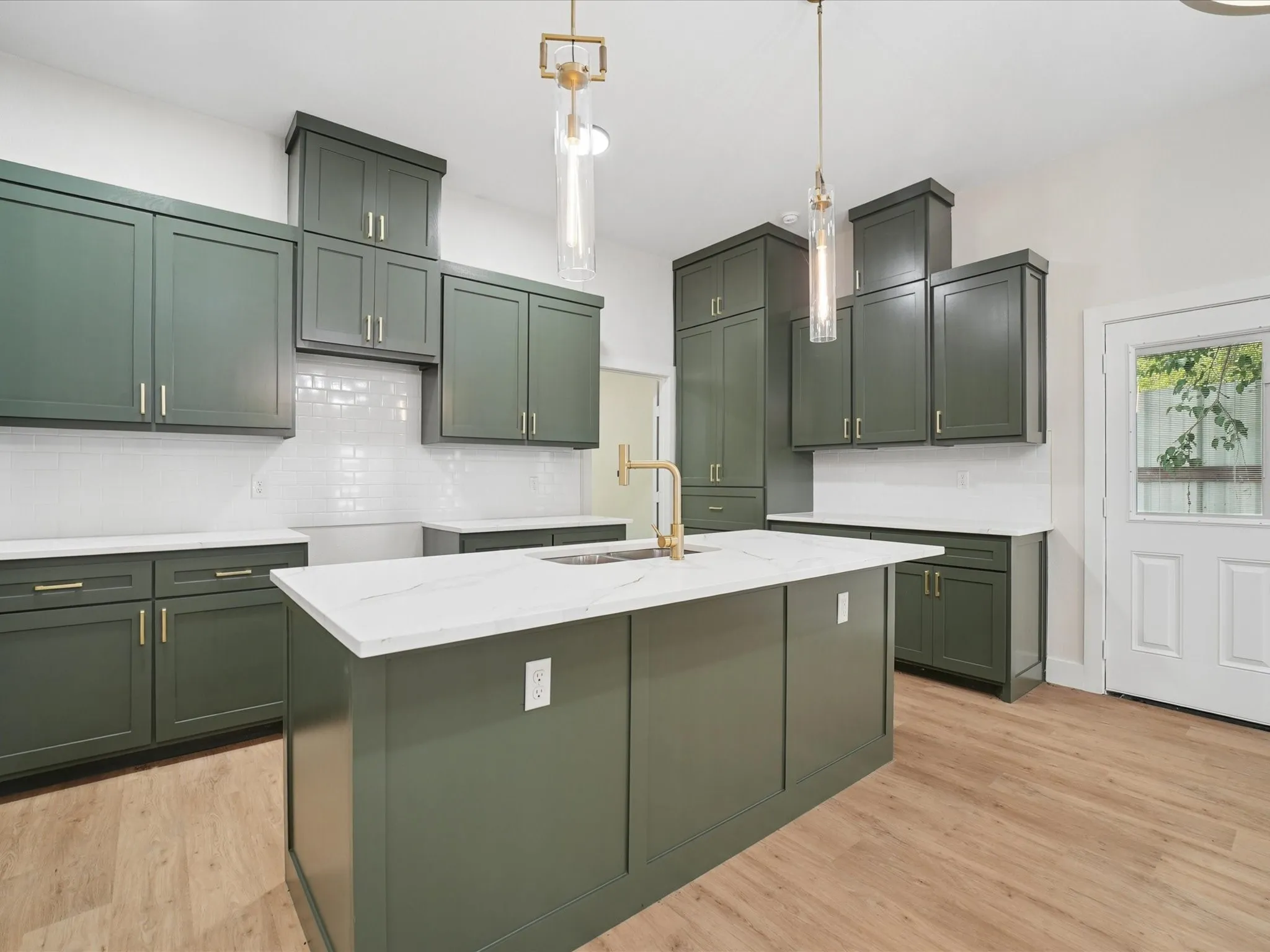Kitchen featuring a central island with an integrated sink, dark green cabinetry, white subway tile backsplash, white countertops, and wood-finish flooring