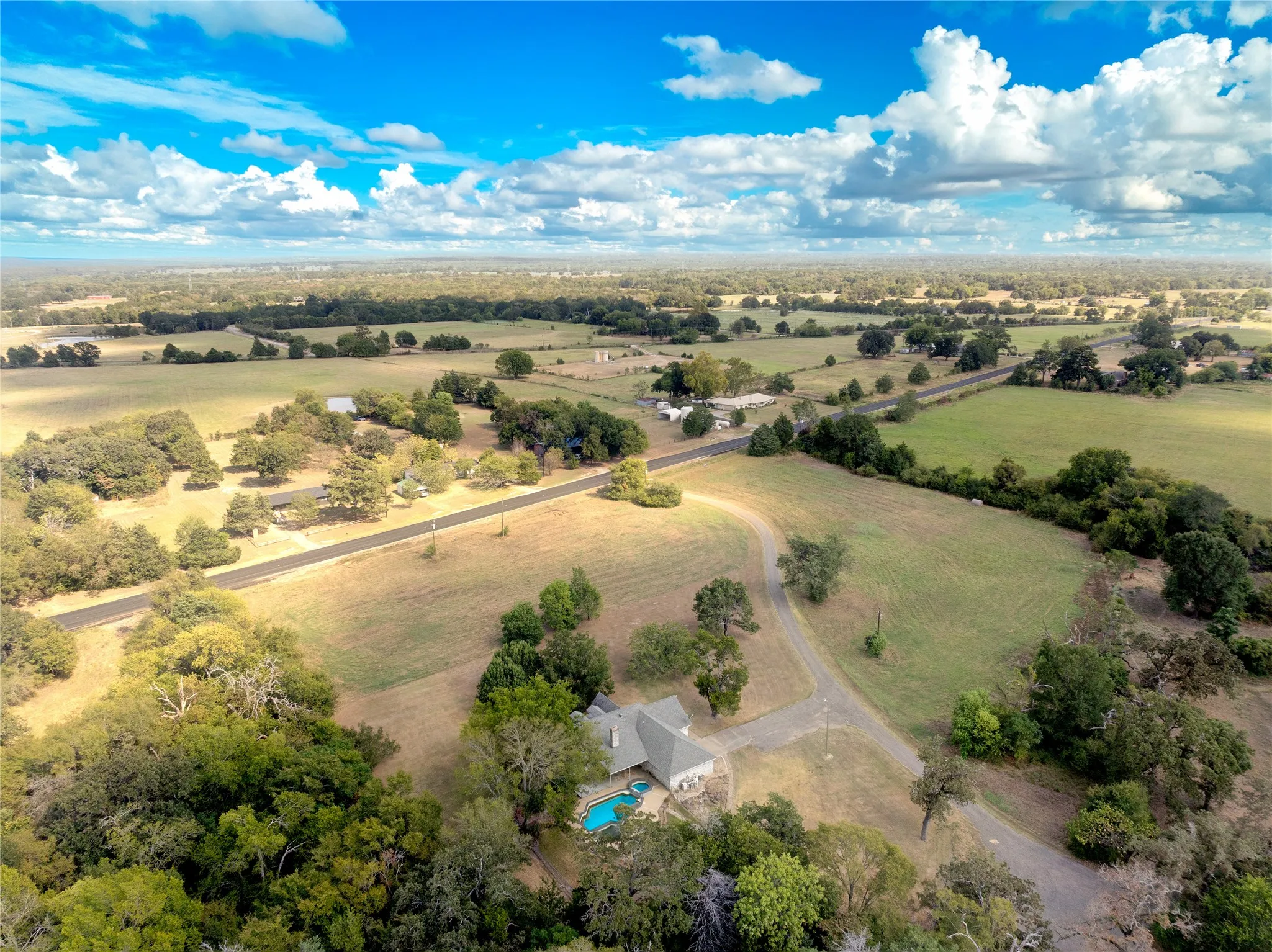 Expansive rural property featuring a single-story residence, a private swimming pool, and a paved driveway, surrounded by mature trees and open fields