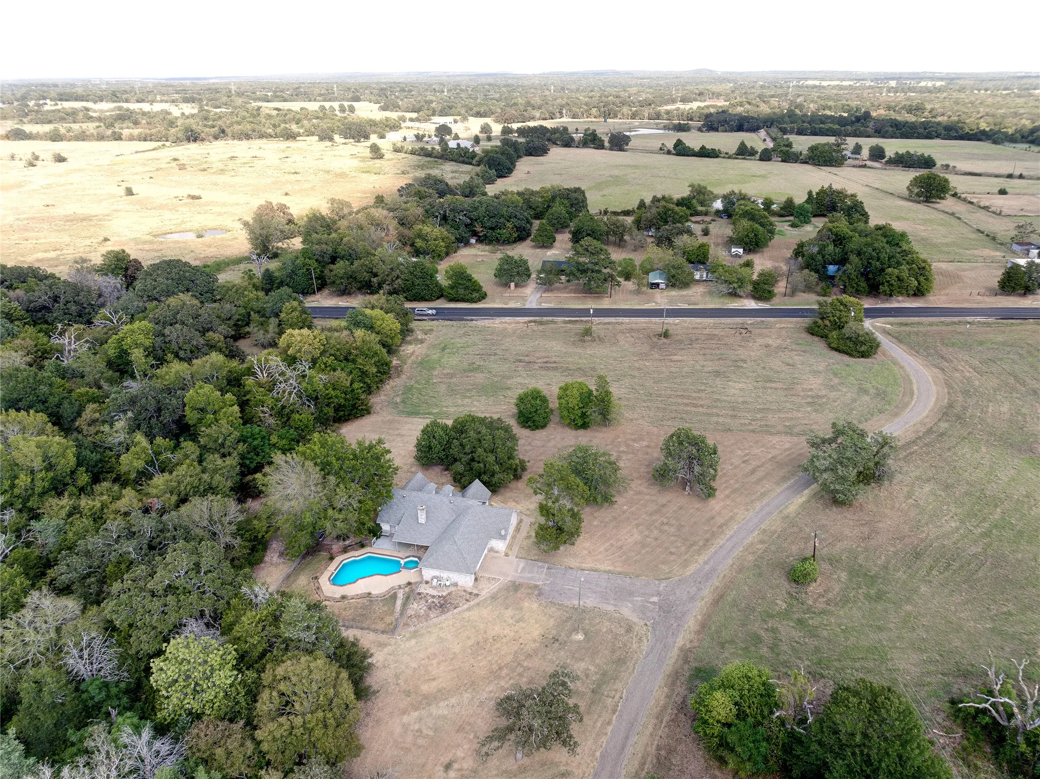 Expansive property featuring a residence, built-in swimming pool, paved driveway, mature trees, and surrounding open land