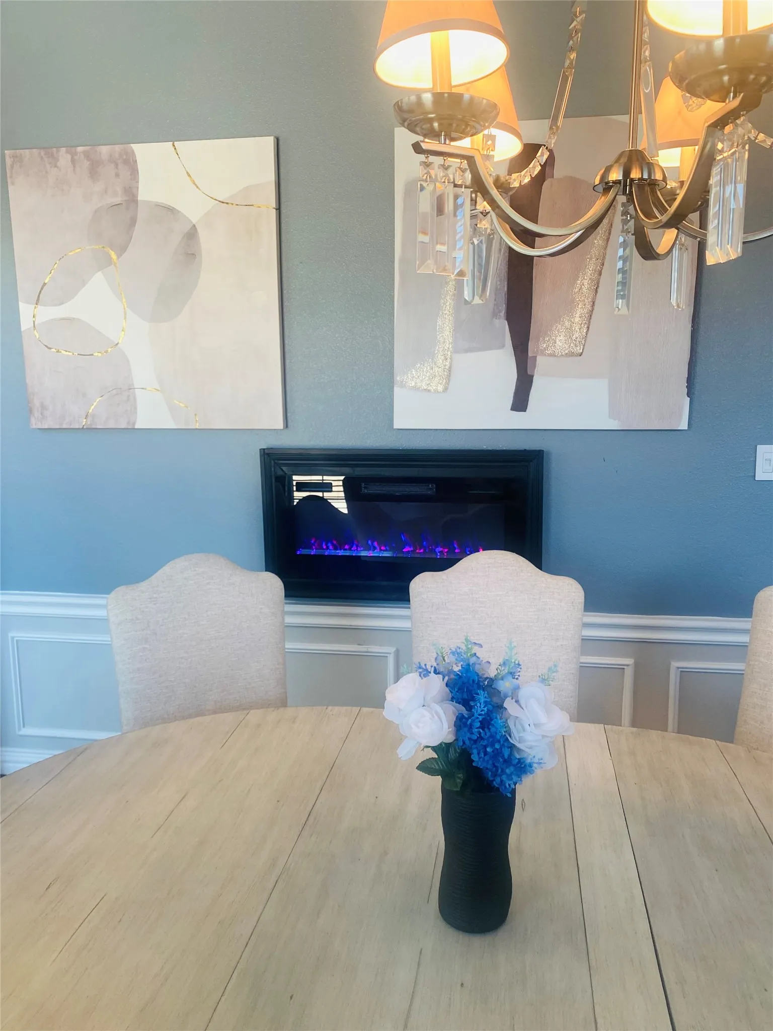Built-in wall fireplace with a dark frame, chandelier with crystal accents and fabric shades, and white wainscoting