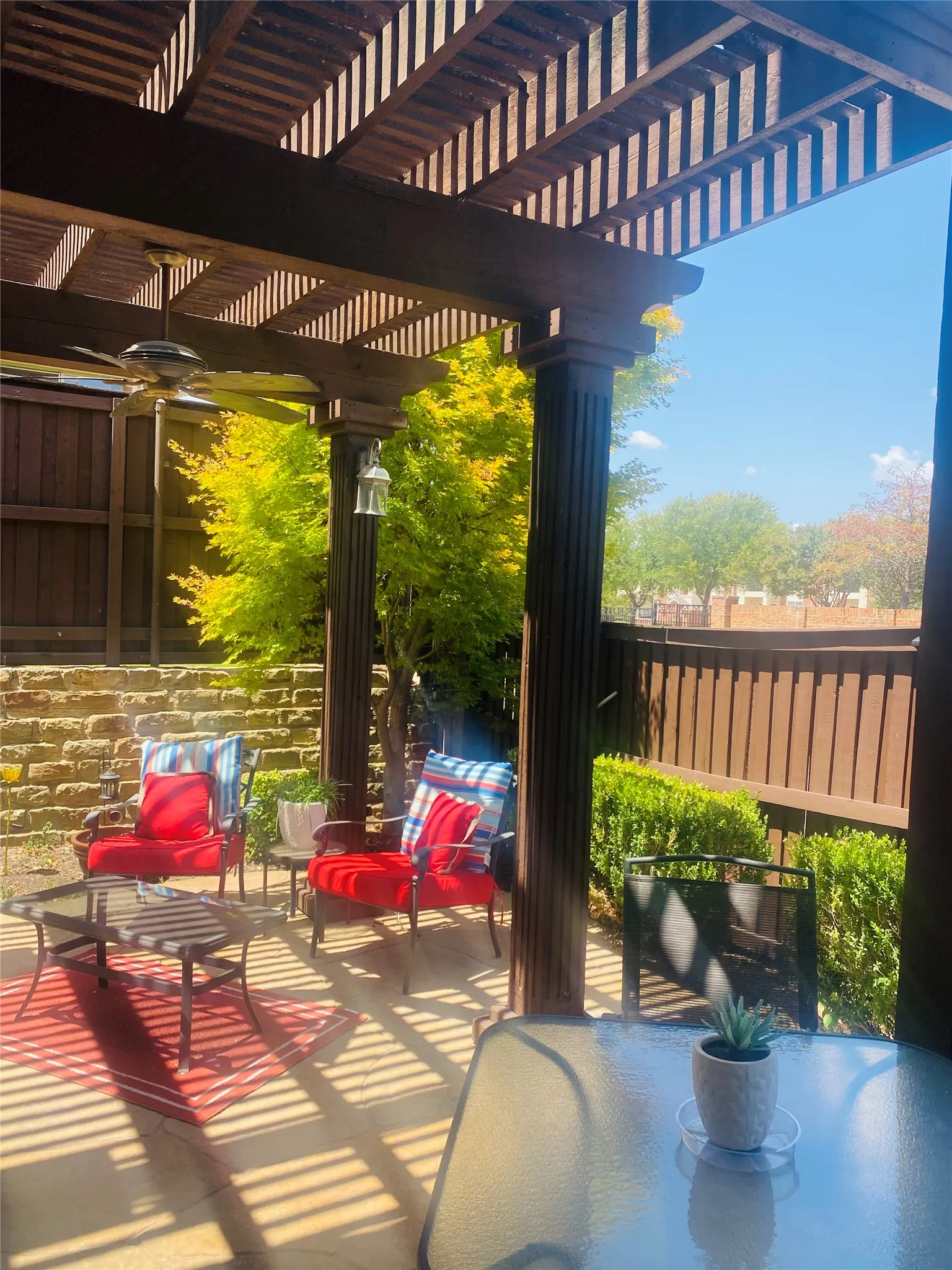Shaded outdoor patio featuring a wood-finish pergola with a ceiling fan, stone-finish retaining wall, and manicured hedges