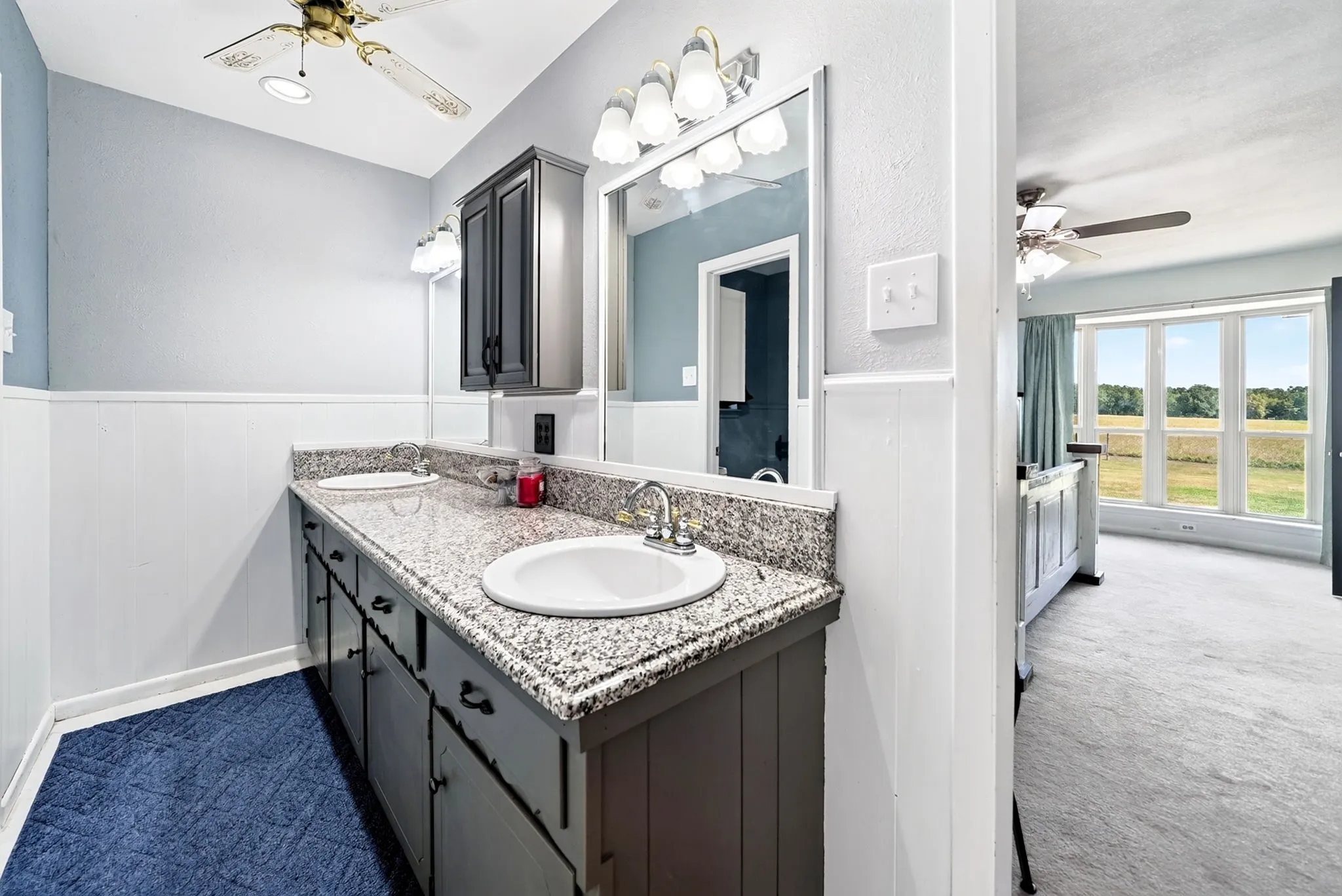 Dual basin vanity with granite-style countertop, white wainscoting, and a ceiling fan with integrated lighting