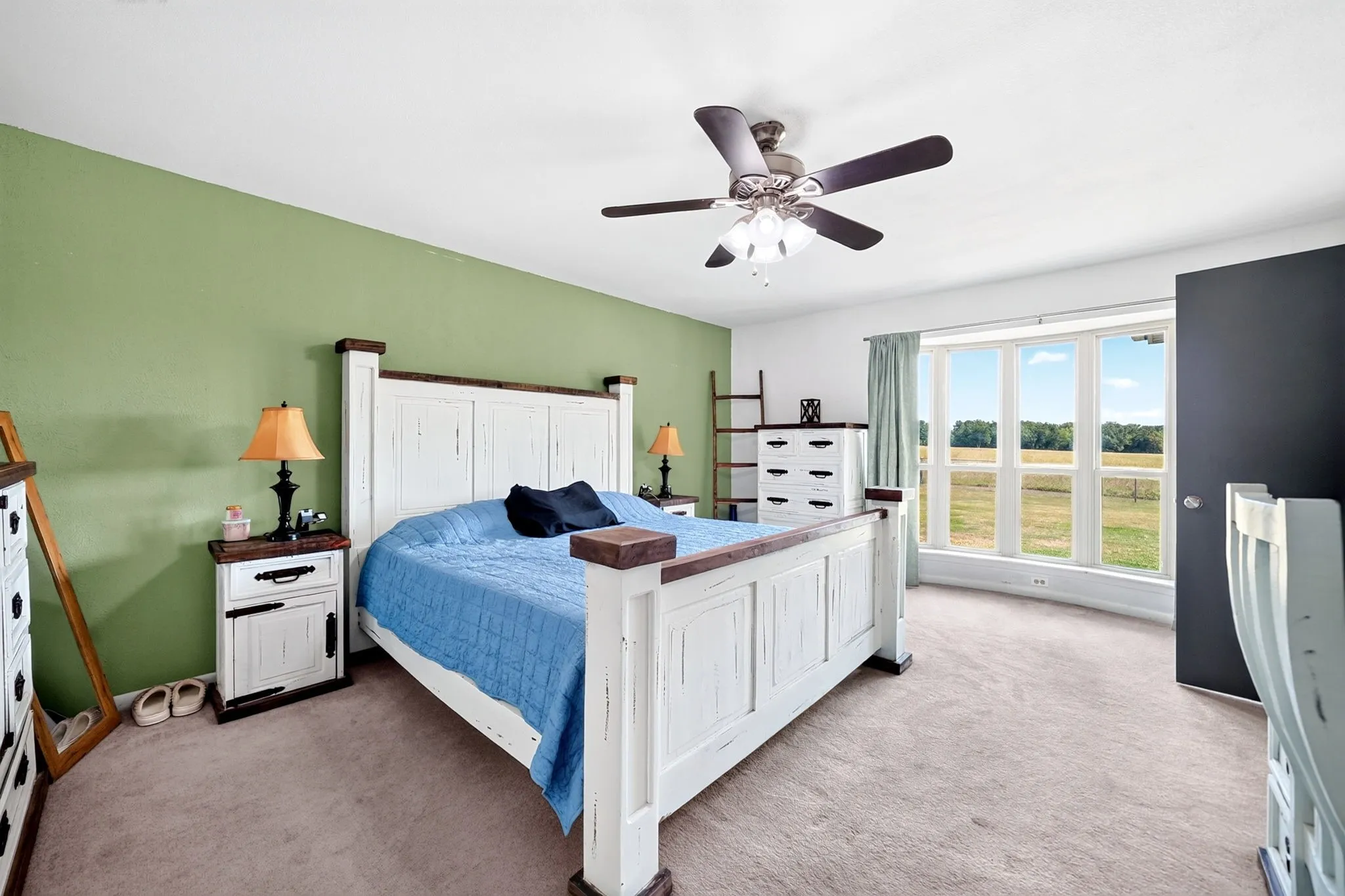 Recessed bay window, ceiling fan with integrated lighting, soft green accent wall, light-toned carpeting, and a dark-paneled door