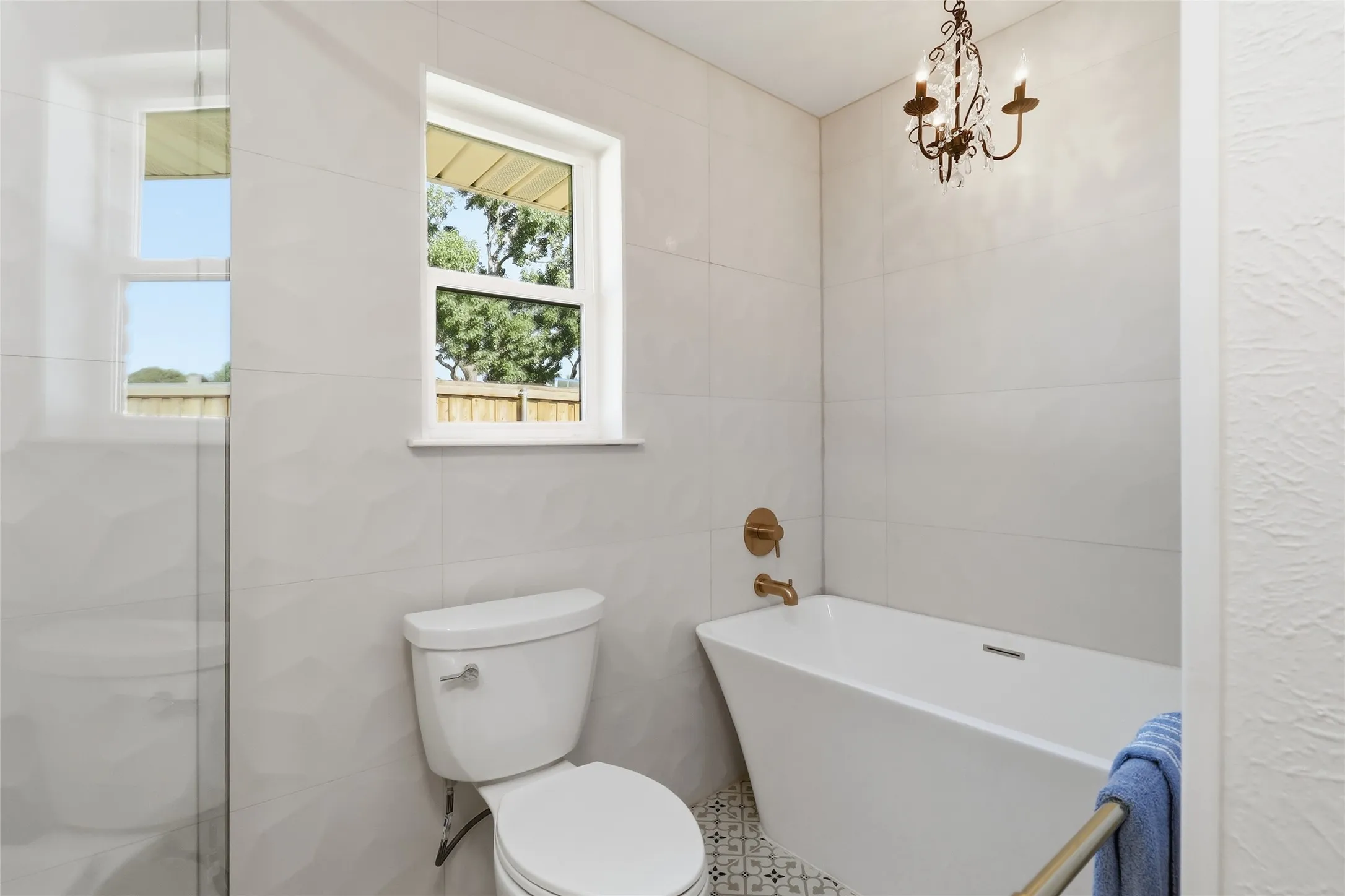 White ceramic wall tiling, free-standing soaking tub, patterned floor tiles, bronze-finish fixtures, and a decorative chandelier