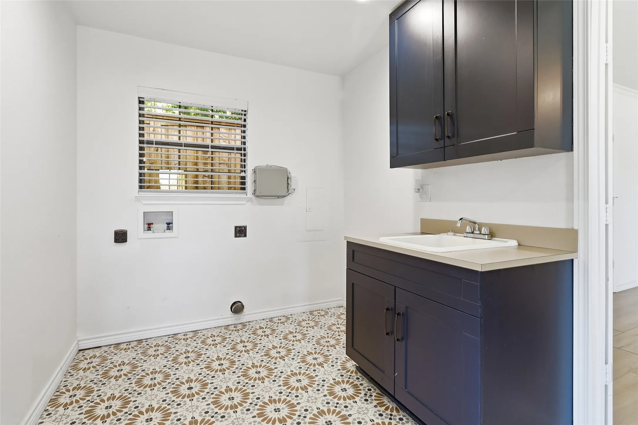 Laundry area with patterned tile flooring, a window with blinds, dark cabinetry, and a utility sink with a quartz countertop