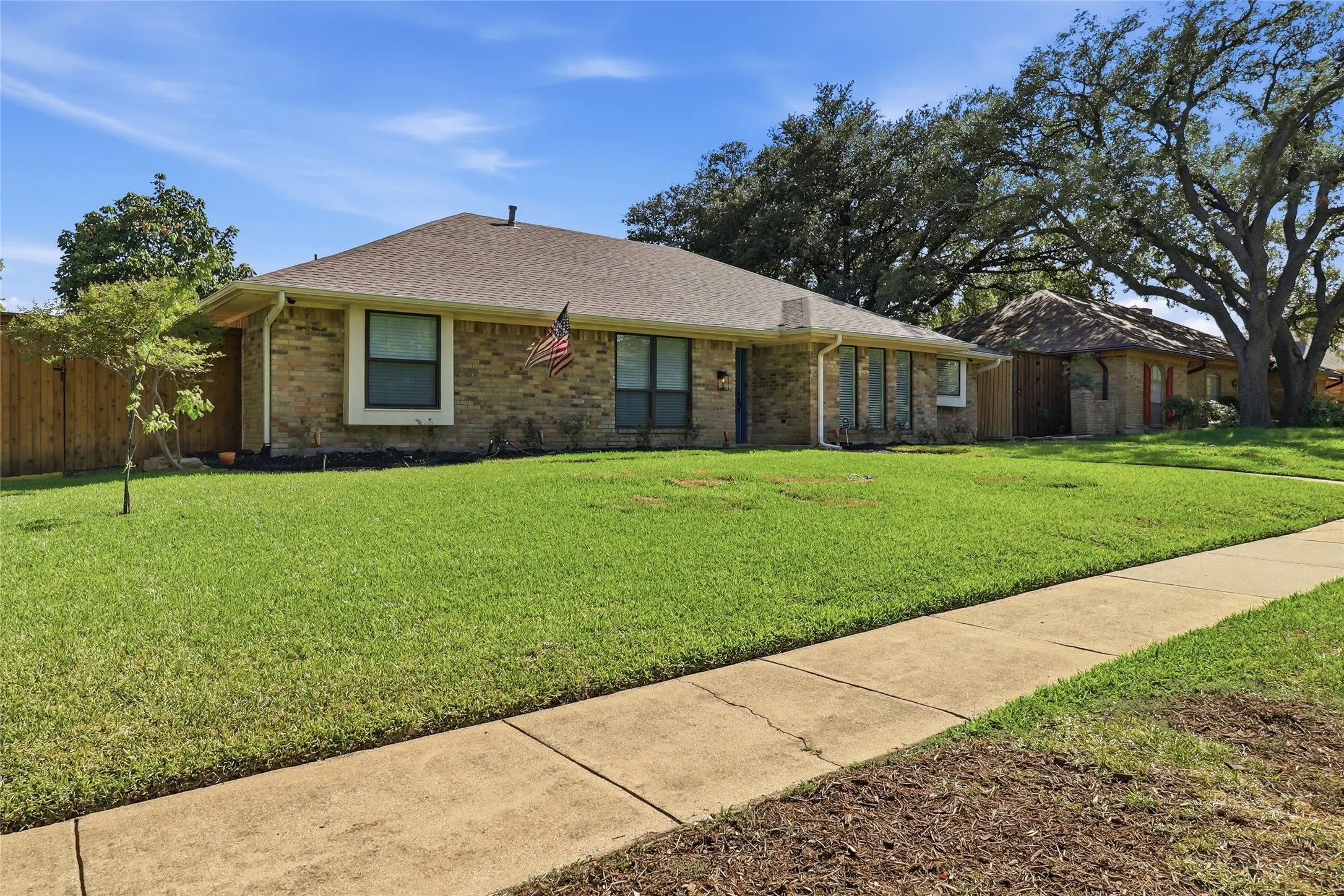 Brick exterior, pitched roof, front yard with mature trees, established landscaping, and concrete sidewalk