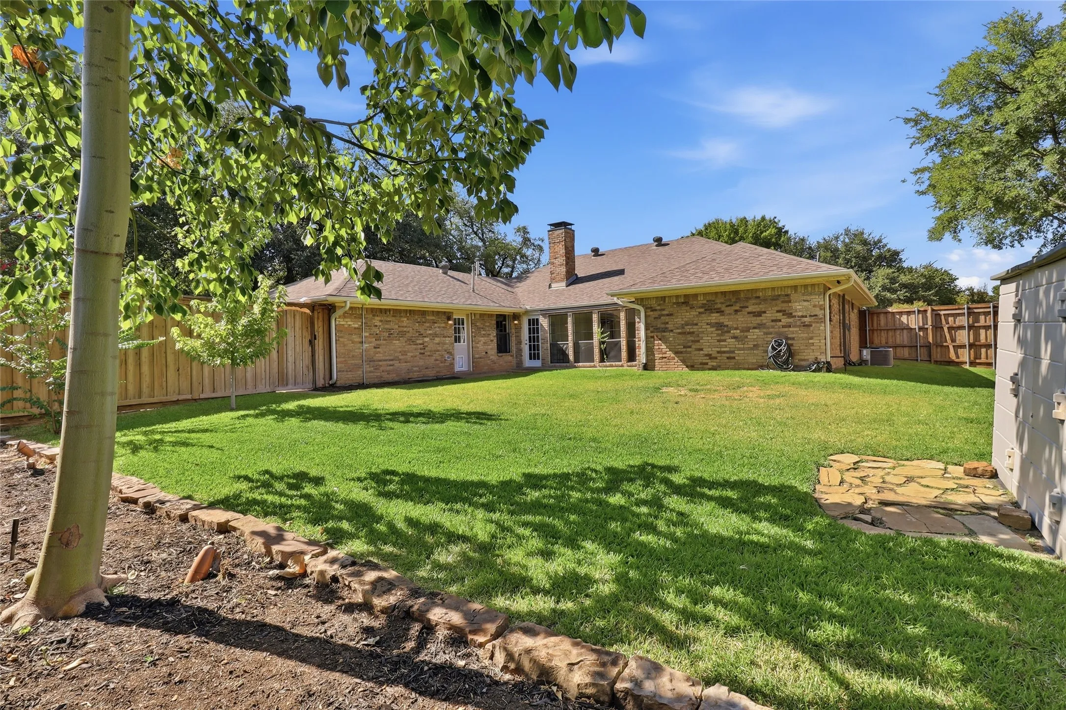 Expansive backyard featuring a manicured lawn, brick exterior, wood fence, and a stone patio
