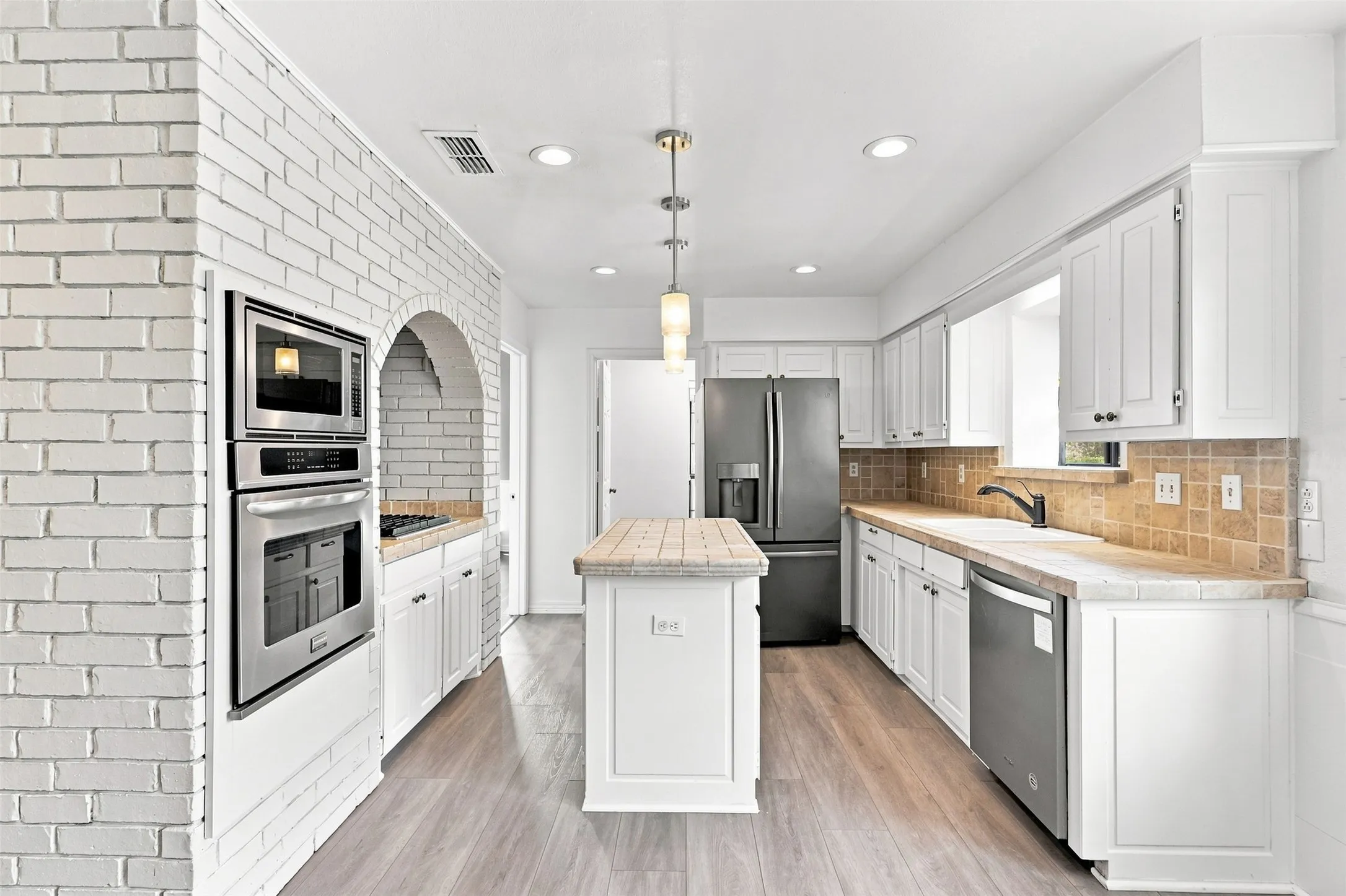 White painted brick archway, built-in stainless steel appliances, wood-finish flooring, white cabinetry, and a center island with a tiled countertop