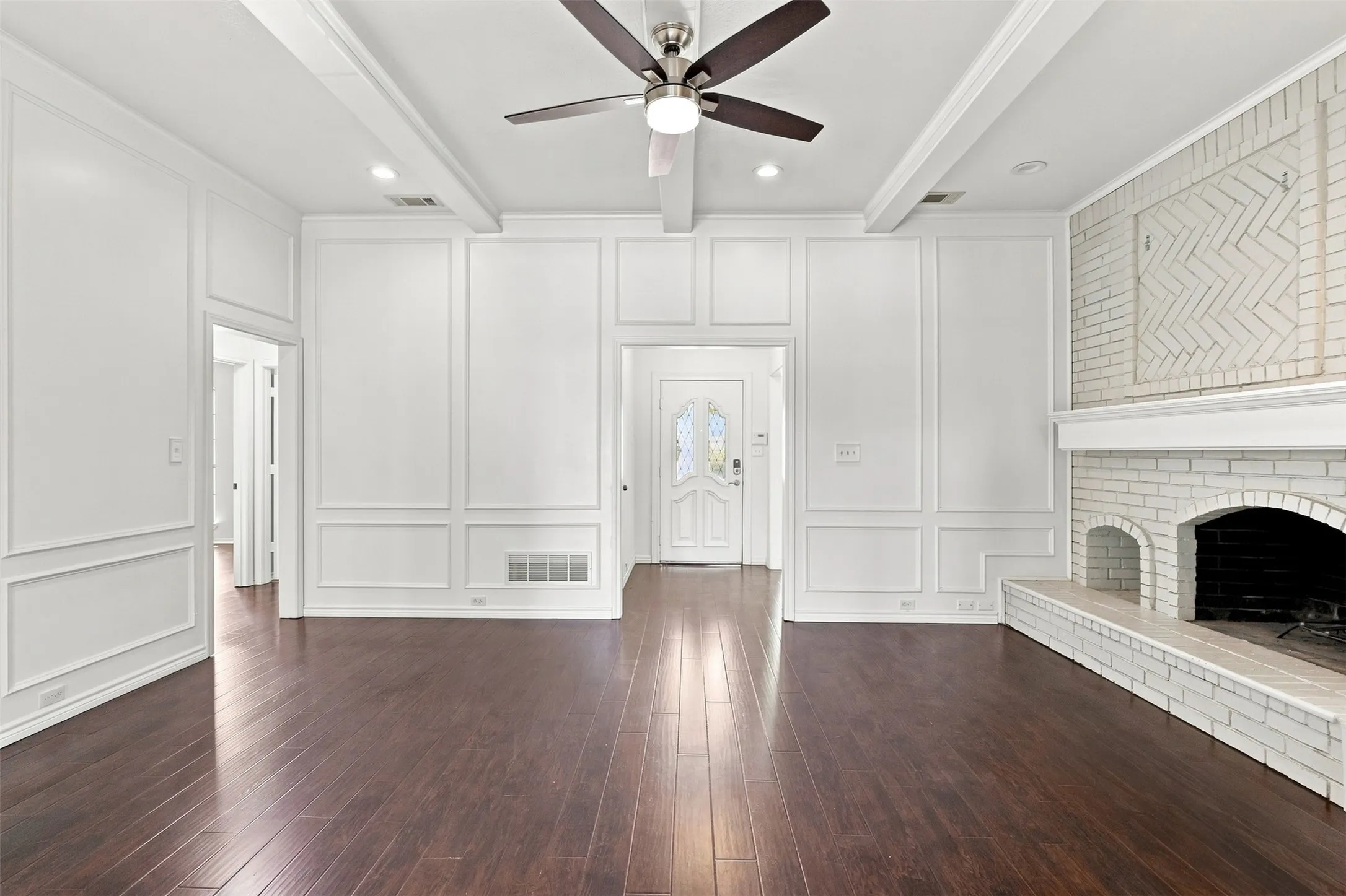 Dark wood-finish flooring, white paneled walls, white brick fireplace with mantel, exposed ceiling beams, and a ceiling fan with integrated lighting