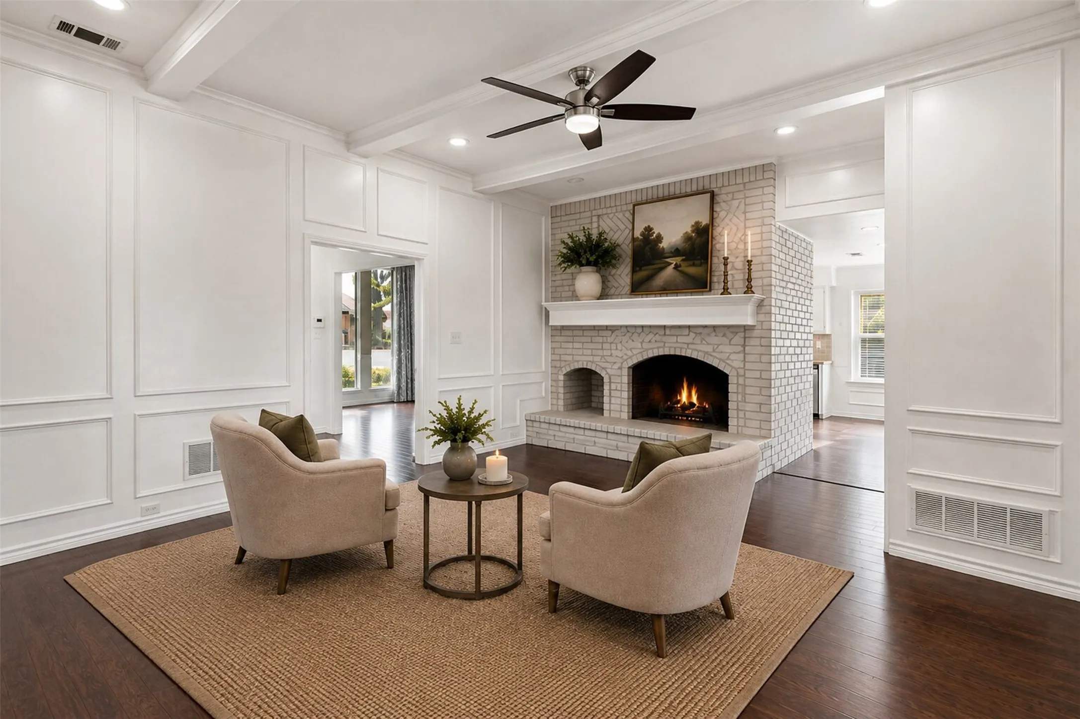 Dark wood-finish flooring, white paneled walls, exposed ceiling beams, recessed lighting, and a white brick fireplace with a decorative mantel