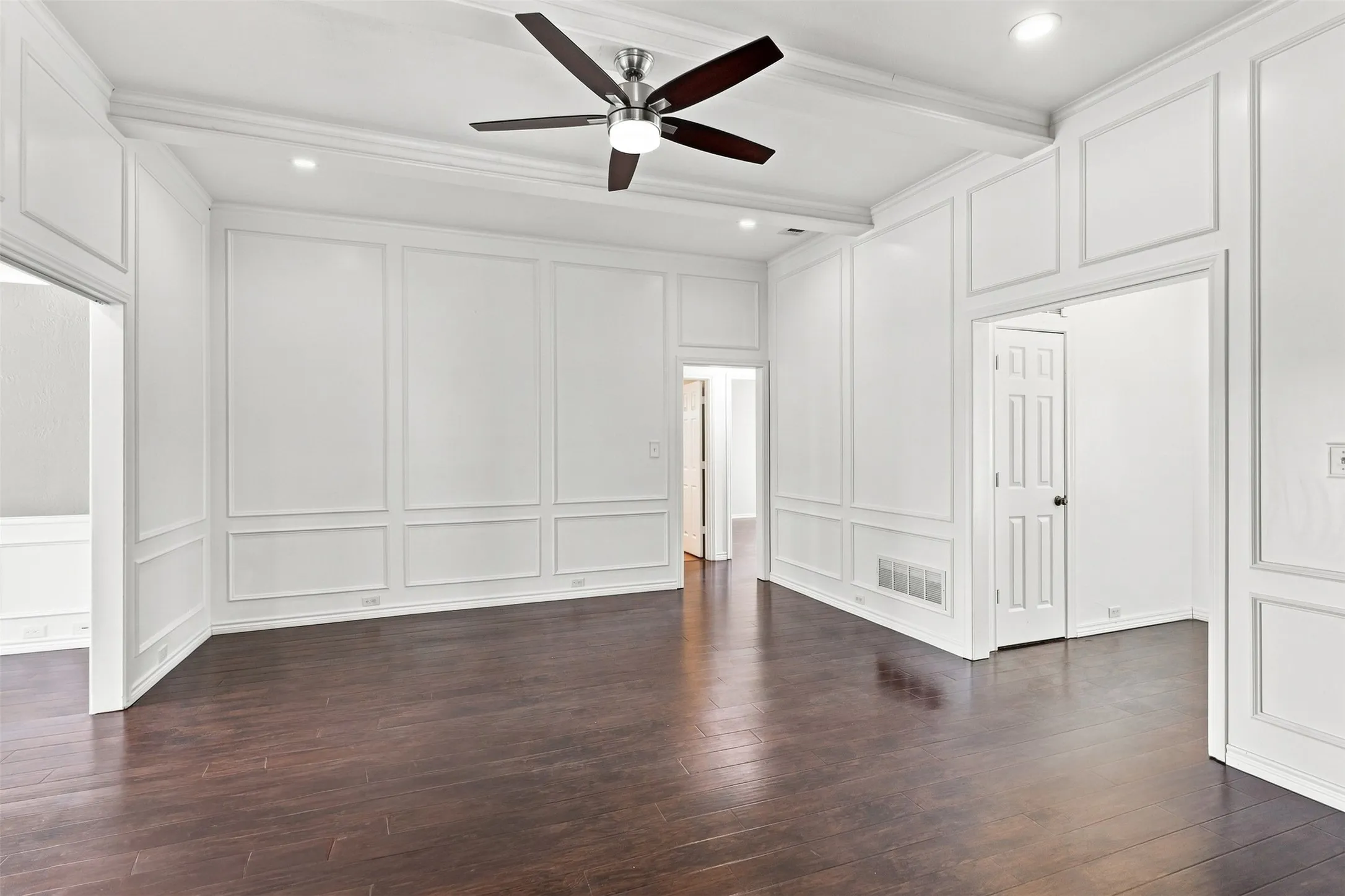 Dark wood-finish flooring, white paneled walls, exposed ceiling beams, recessed lighting, and a white brick fireplace with a decorative mantel