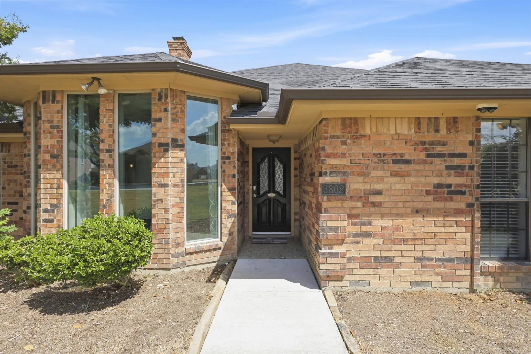 Brick exterior with a prominent bay window, dark shingle roof, and a black front door with decorative glass
