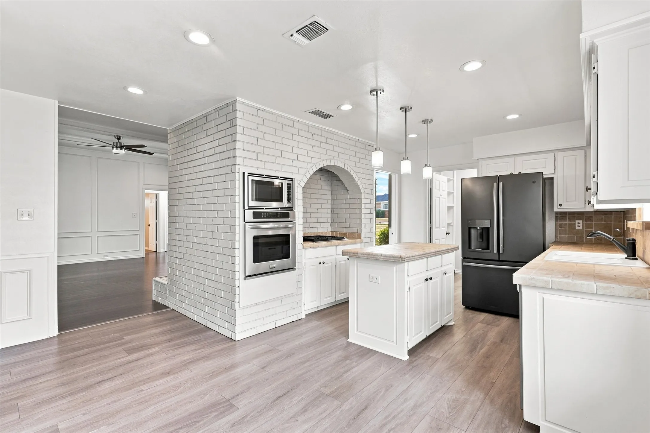 Wood-finish flooring throughout, white brick archway, stainless steel built-in appliances, white cabinetry, and a kitchen island with pendant lighting