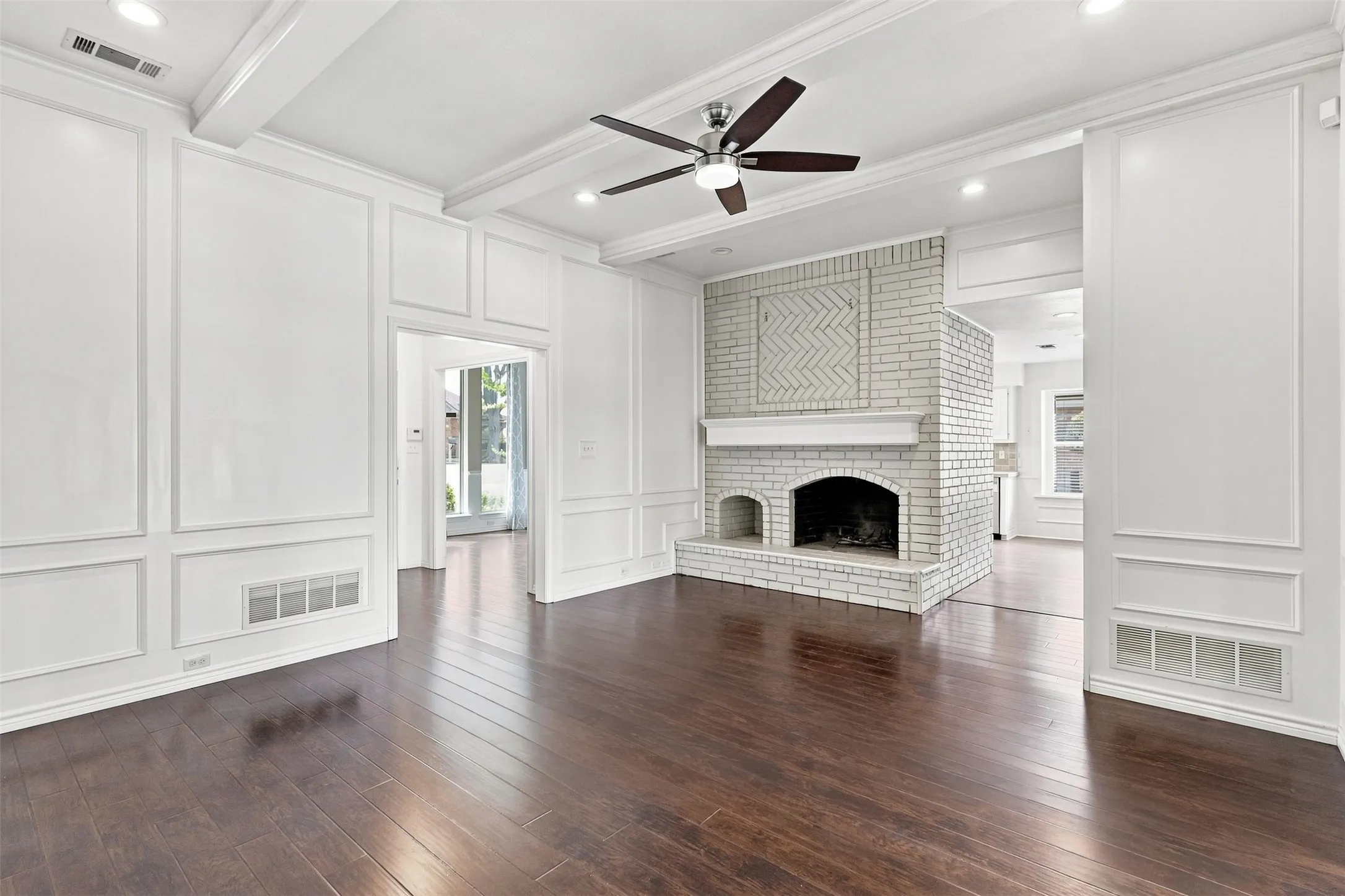Dark wood-finish flooring, white paneled walls, exposed ceiling beams, recessed lighting, and a white brick fireplace with a decorative mantel