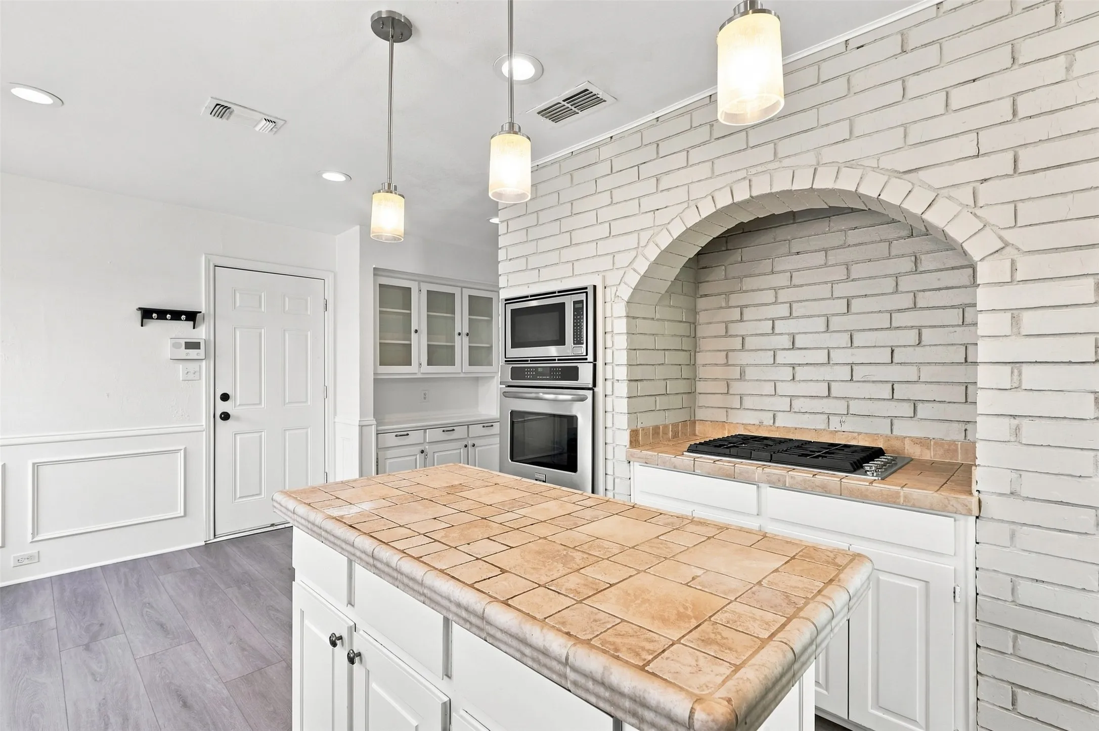 Kitchen featuring a tiled island, built-in oven and microwave, white brick archway, paneled wainscoting, and wood-finish flooring