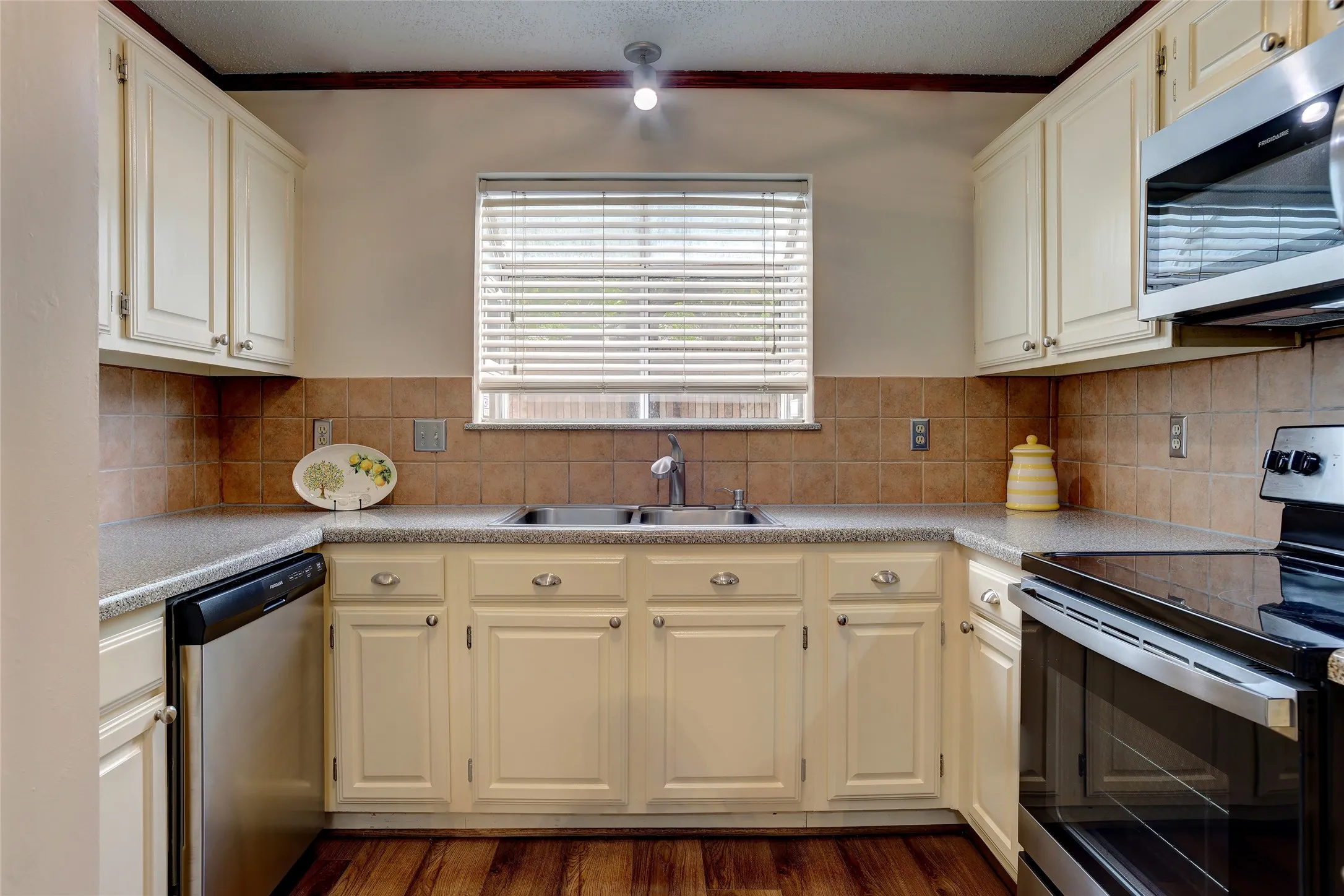 Galley kitchen featuring cream-painted cabinetry with raised-panel doors, wood-finish flooring, tile backsplash, and stainless steel appliances.