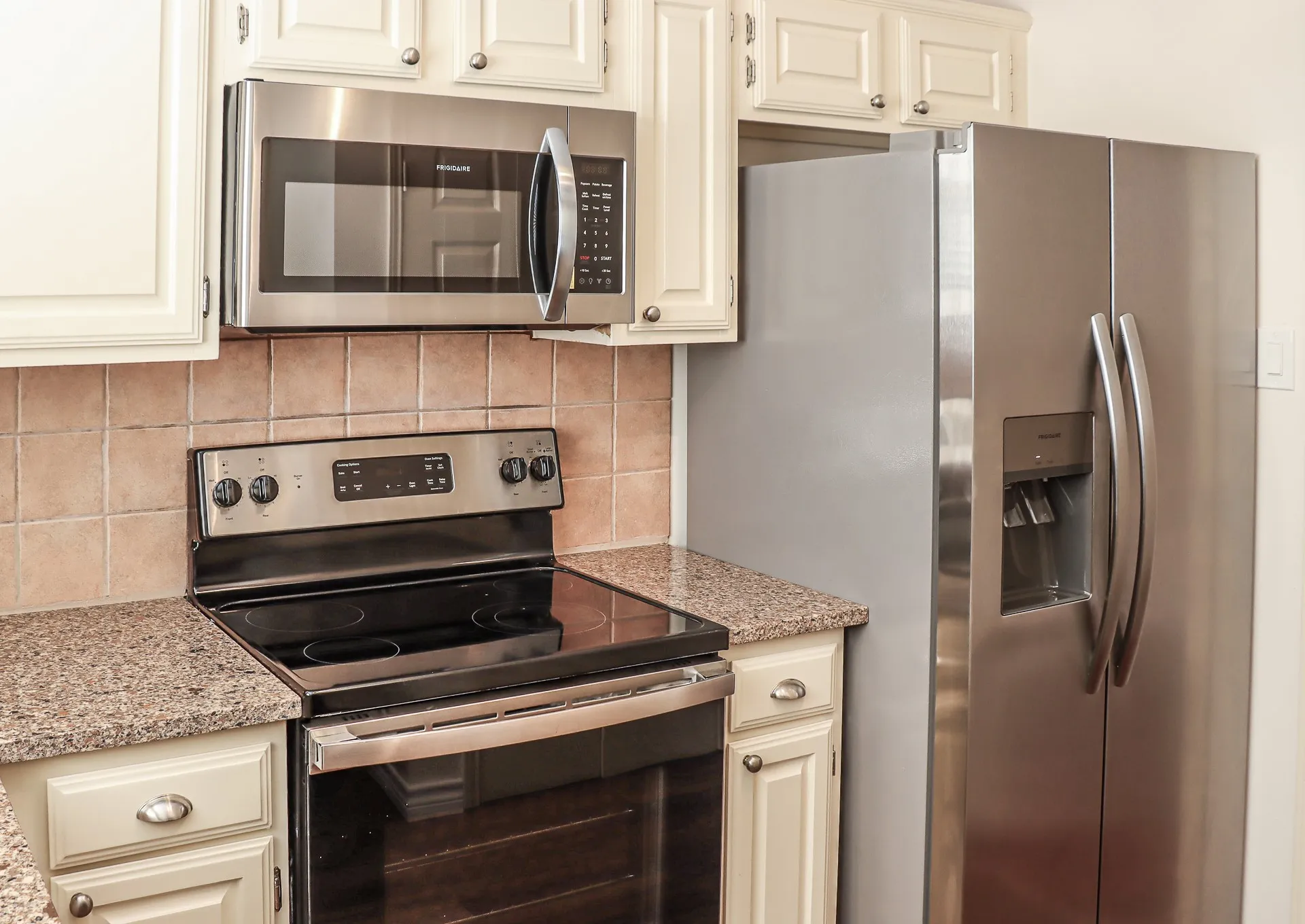 Updated kitchen featuring cream-colored cabinetry, granite countertops, stainless steel appliances including a side-by-side refrigerator with ice and water dispensers, electric range with cooktop, and overhead microwave.