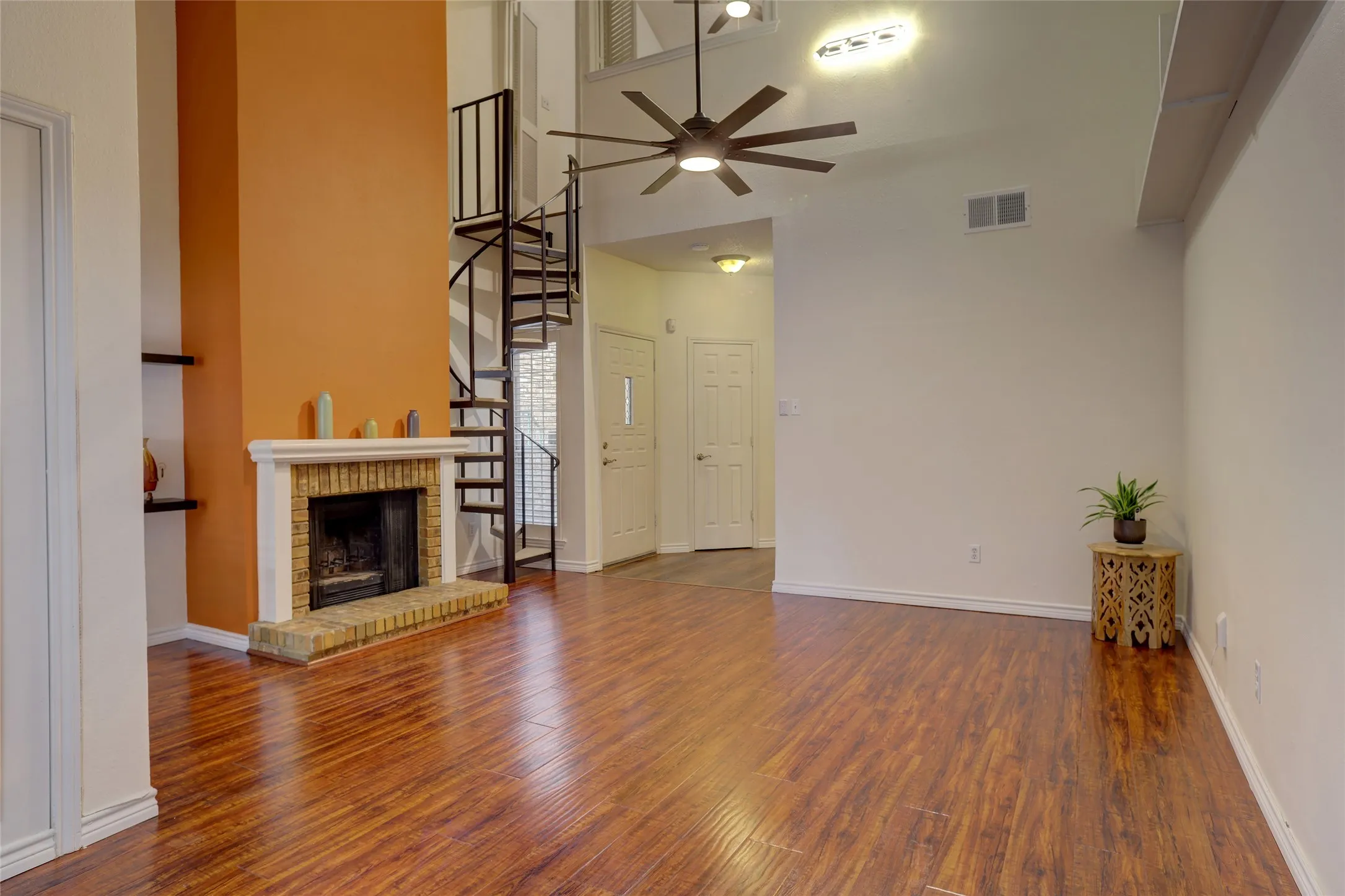 Open-concept living space featuring warm wood-finish flooring throughout, a decorative fireplace with brick surround, modern ceiling fan with integrated lighting, and bright, neutral wall finishes