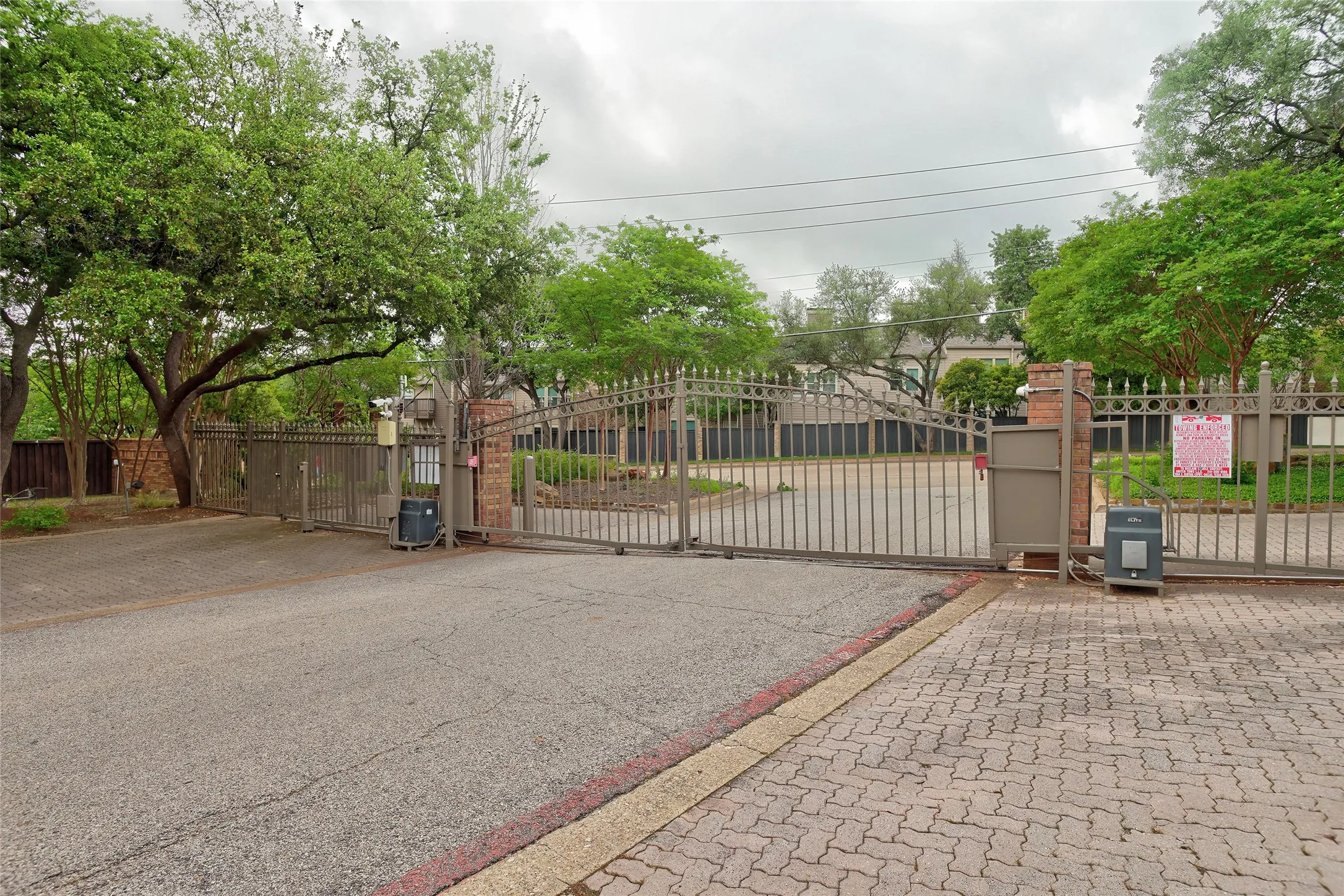 Secured entry featuring wrought-iron gates with brick and stone pillars, complemented by mature shade trees and brick paver hardscaping throughout the grounds