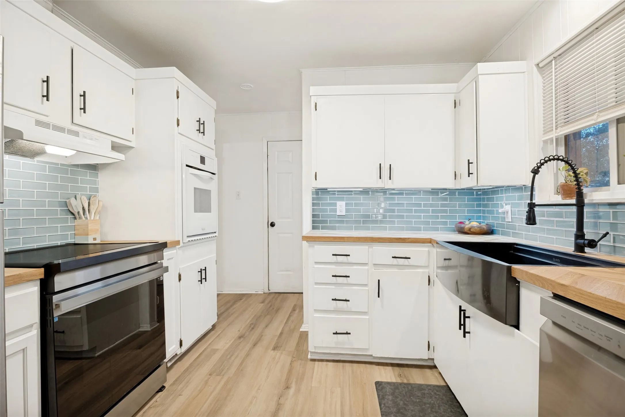 White cabinetry with matte black hardware, wood-finish countertops, stainless steel appliances, blue subway tile backsplash, and wood-finish flooring