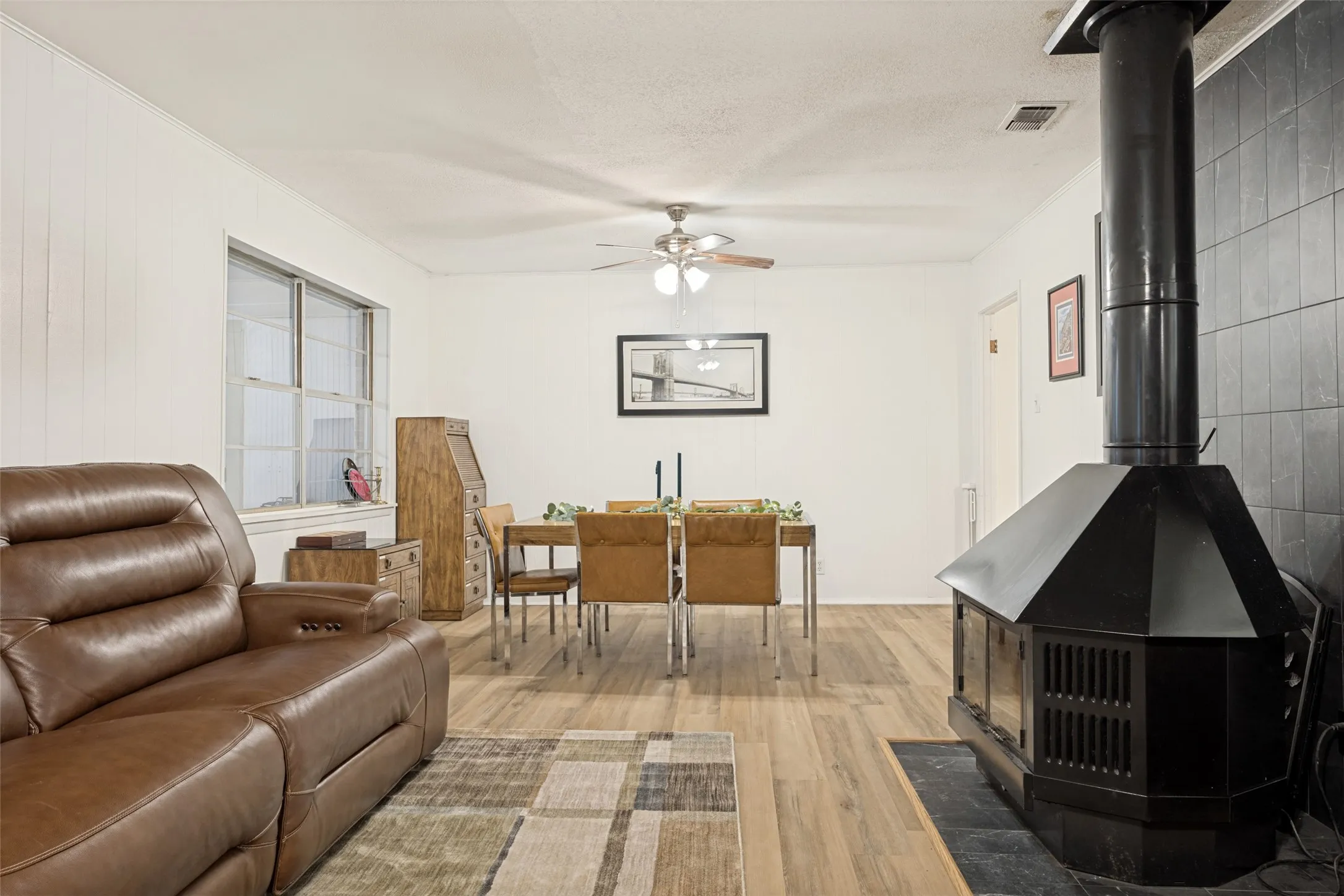 Open-concept living area featuring wood-finish flooring, a ceiling fan with integrated lighting, and a black metal wood-burning fireplace with a tiled surround