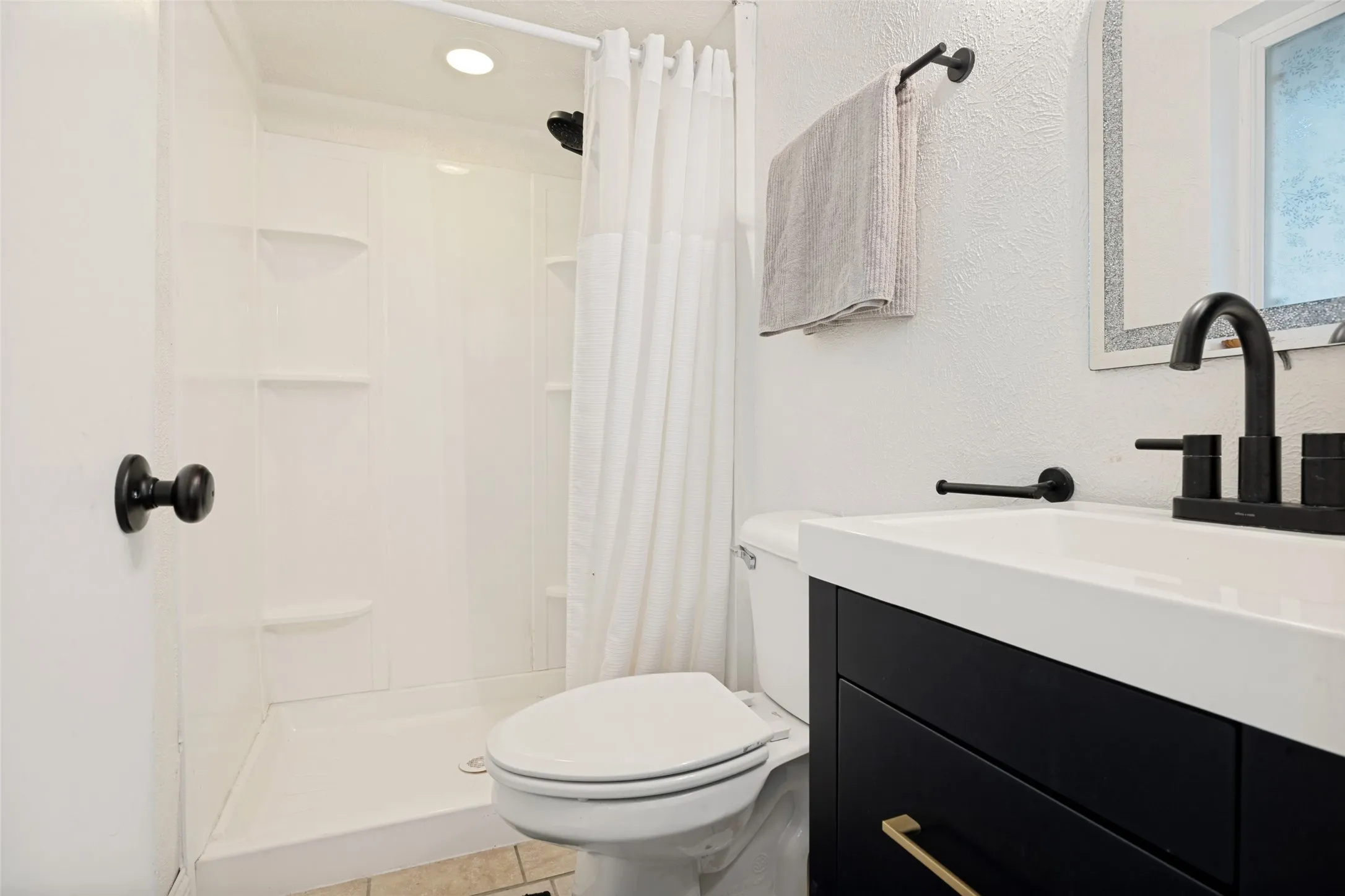 White shower enclosure with built-in shelving, matte black fixtures, dark wood-finish vanity with gold hardware, and a white integrated sink