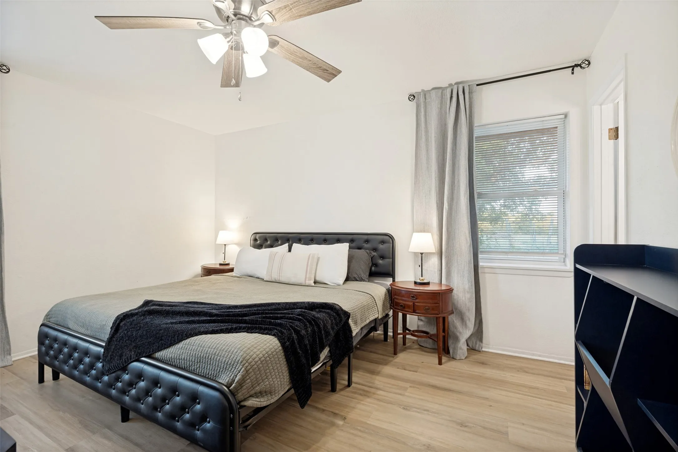 Light wood-finish flooring, a ceiling fan with integrated lighting, and a window with blinds provide comfort