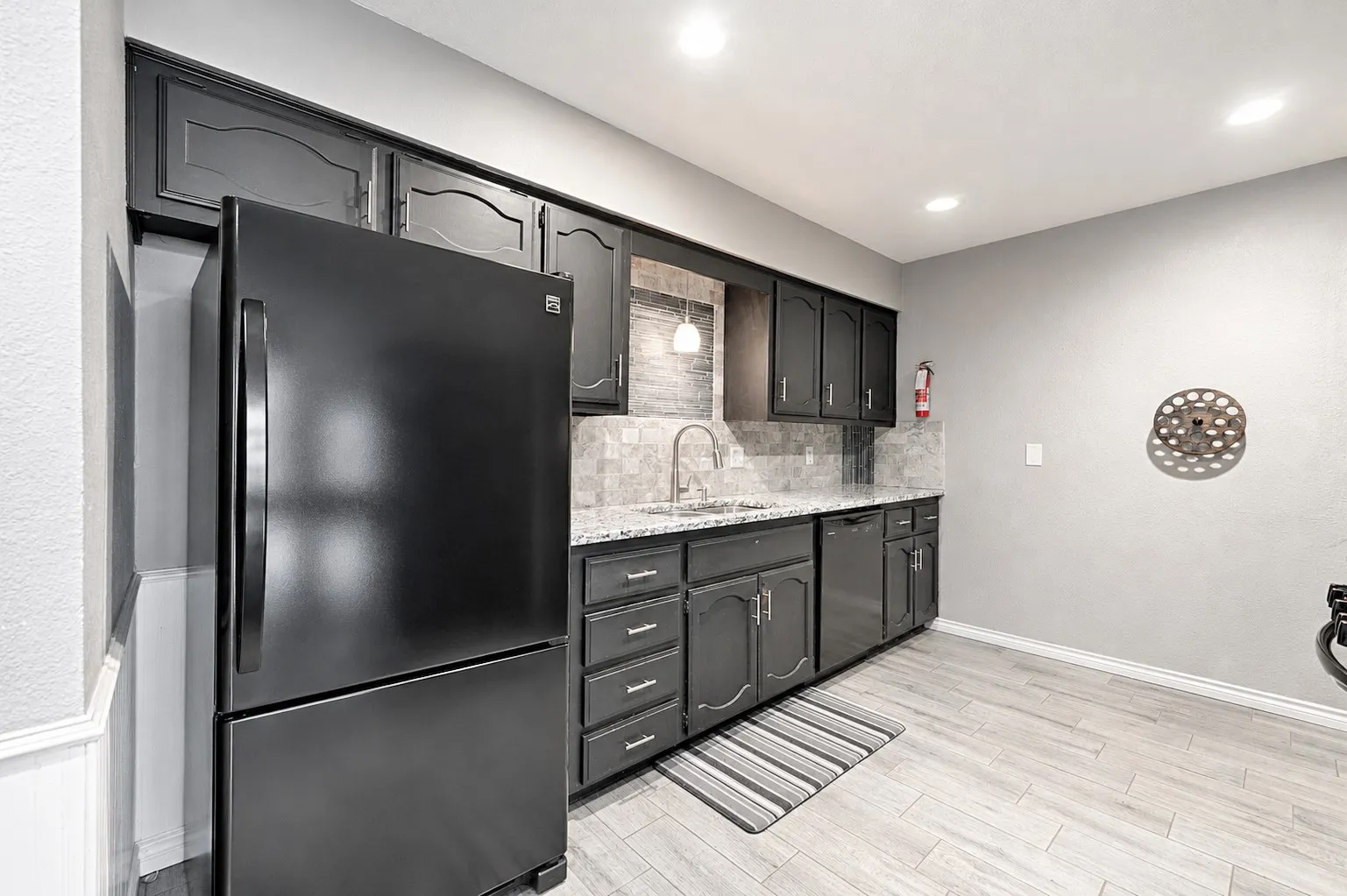 Black cabinetry with decorative molding, light-toned tile backsplash, granite-style countertops, integrated stainless-steel sink, and wood-finish flooring