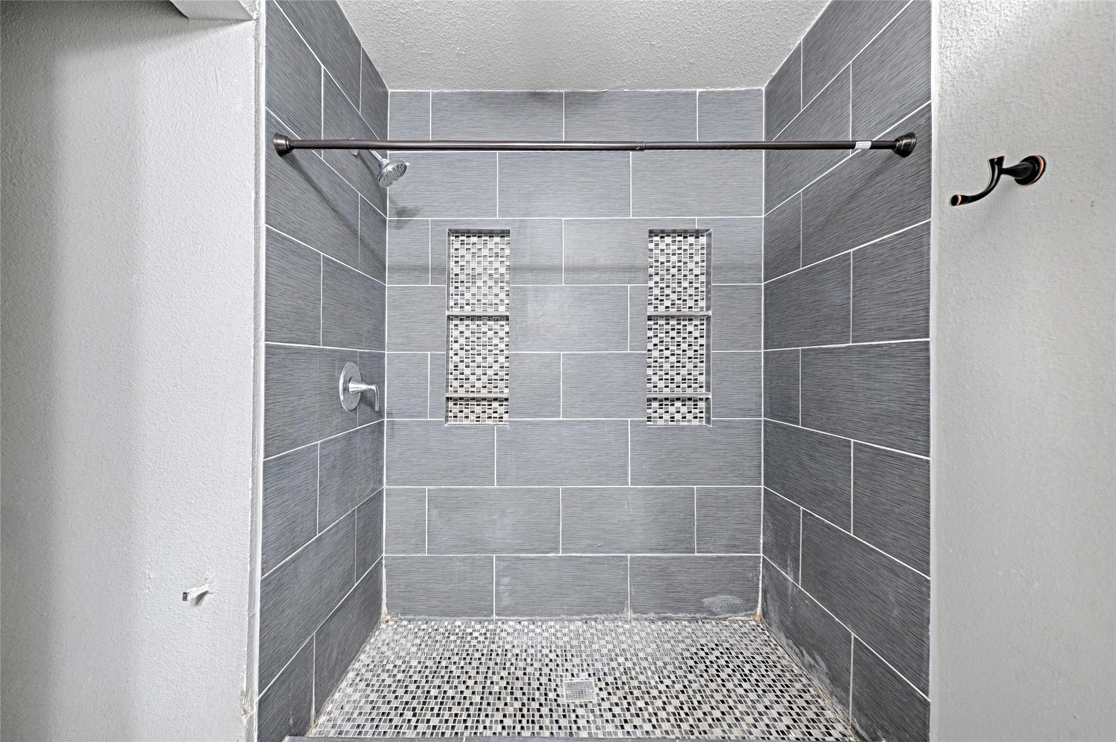Tiled shower enclosure featuring rectangular gray wall tiles, mosaic tile shower floor, and two recessed mosaic tile niches