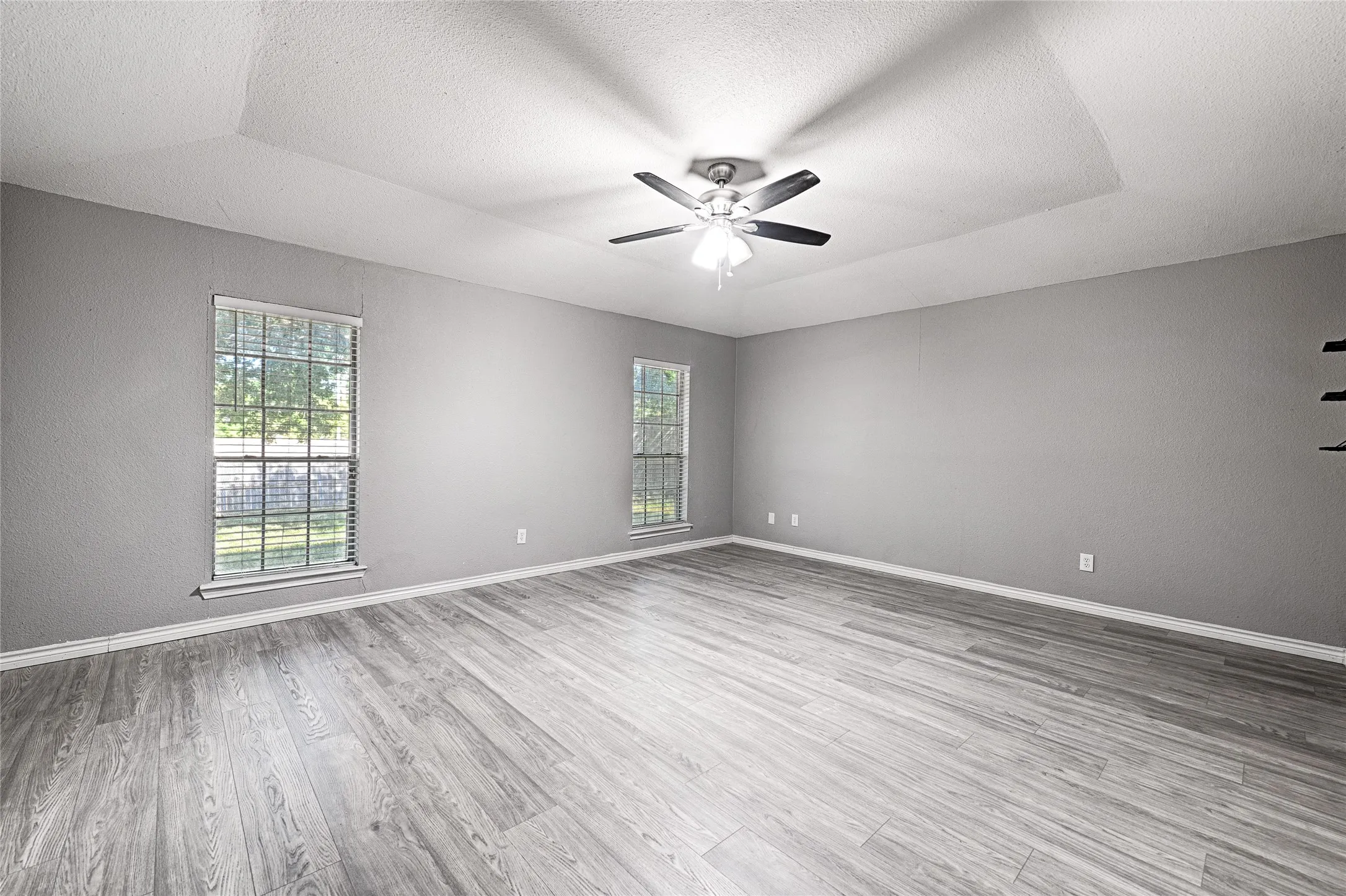 Expansive room featuring wood-finish flooring, two windows with blinds, a tray ceiling, and a ceiling fan with light fixture