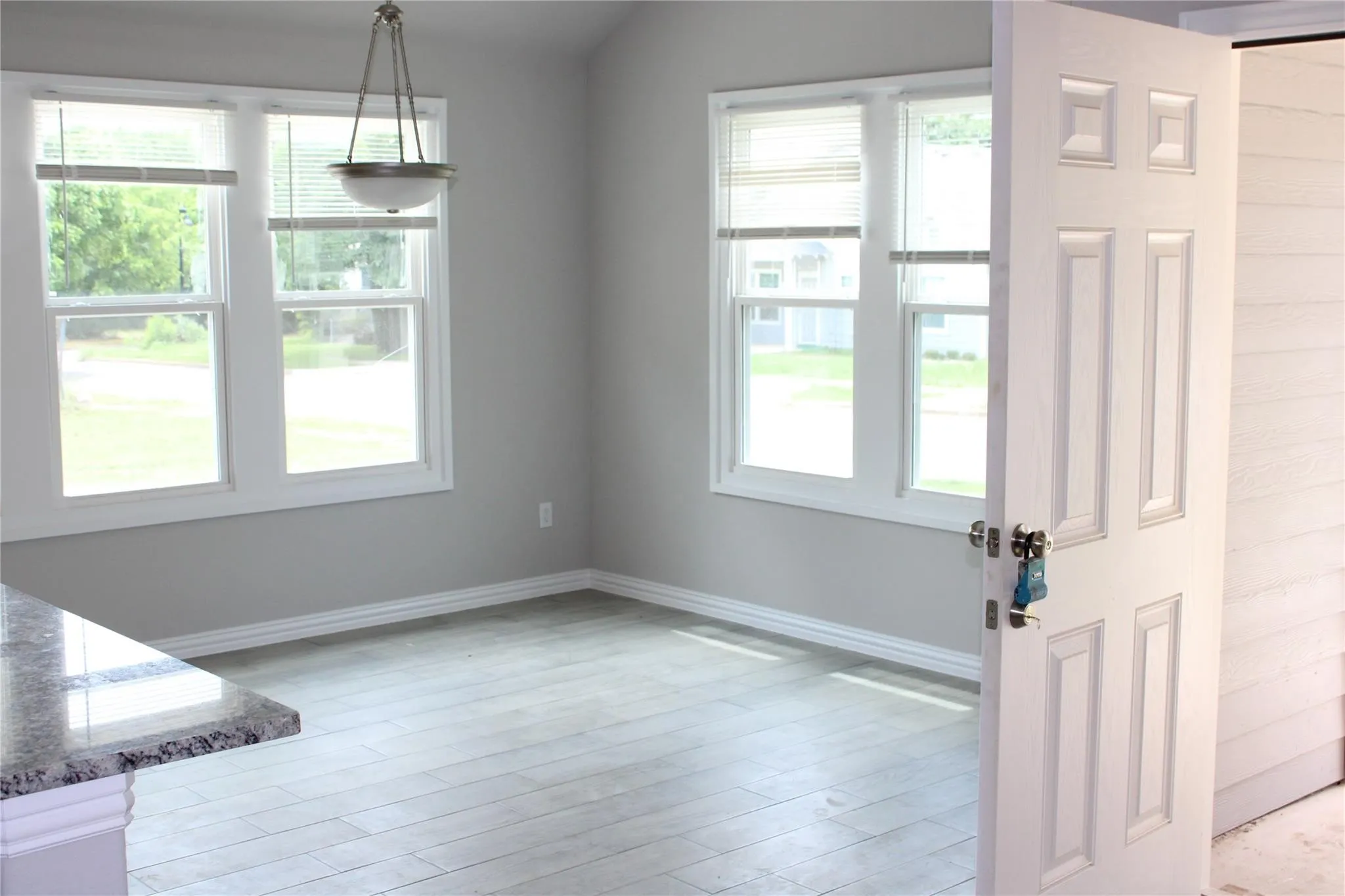 Light gray walls with white trim, wood-finish flooring, two large windows with blinds, a hanging light fixture, and a granite countertop