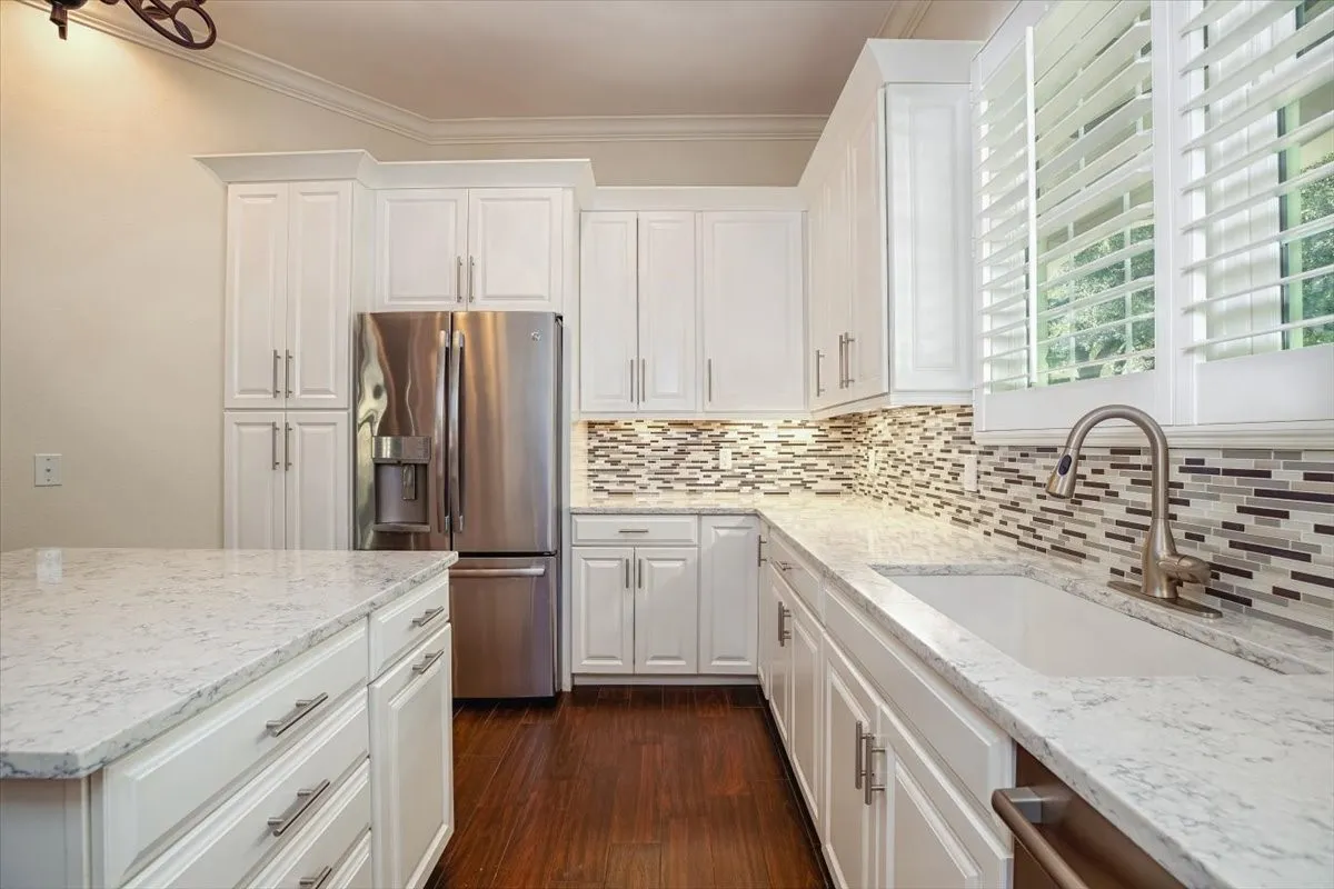 White cabinetry with brushed nickel hardware, light-toned countertops, mosaic tile backsplash, stainless steel refrigerator, and porcelain wood-finish flooring