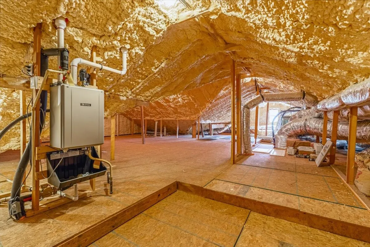 Attic space featuring spray foam insulation, exposed wooden beams, an installed tankless water heater, and ductwork