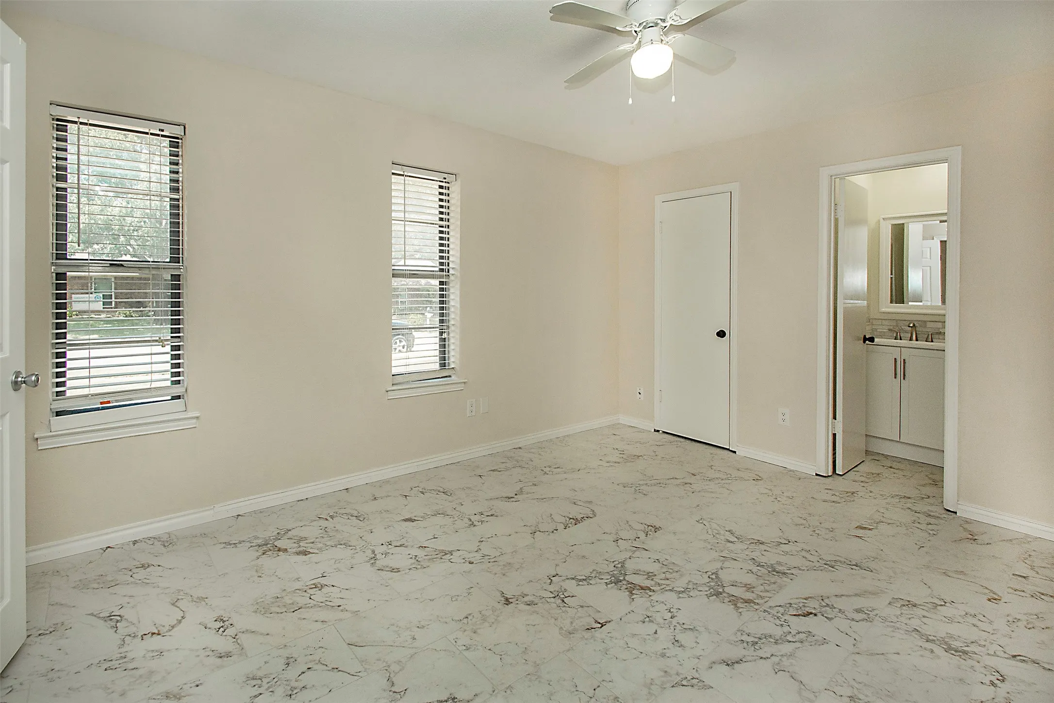 Spacious room featuring patterned flooring, two windows with blinds, a ceiling fan with integrated lighting, and an attached bathroom with a single vanity