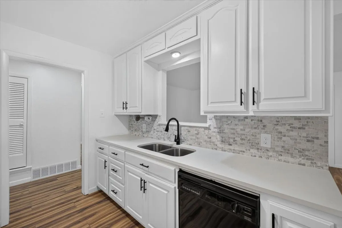 White cabinetry with black hardware, stone-finish countertops, stainless steel double basin sink, mosaic tile backsplash, and wood-finish flooring
