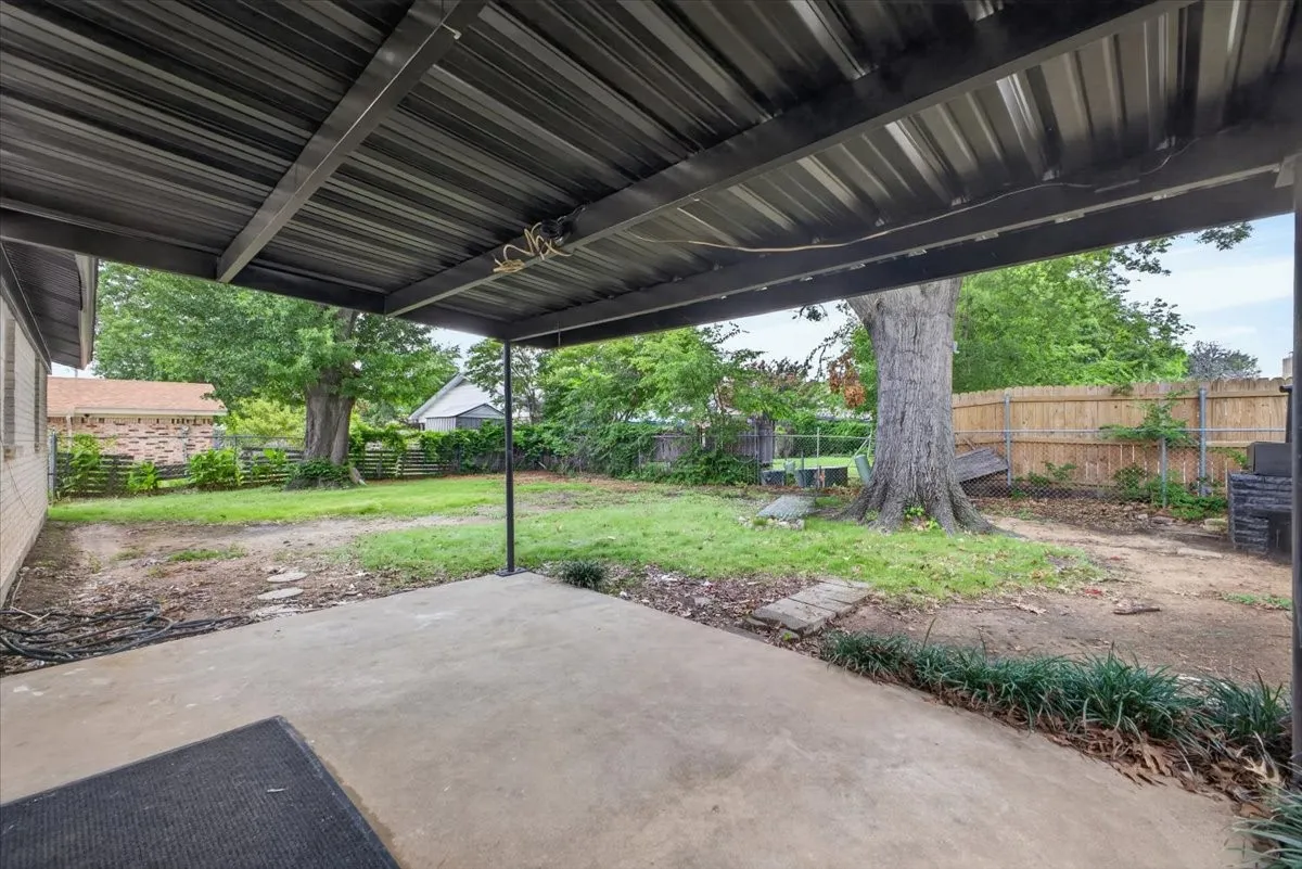 Covered concrete patio with a metal roof structure, overlooking a fenced backyard with mature trees