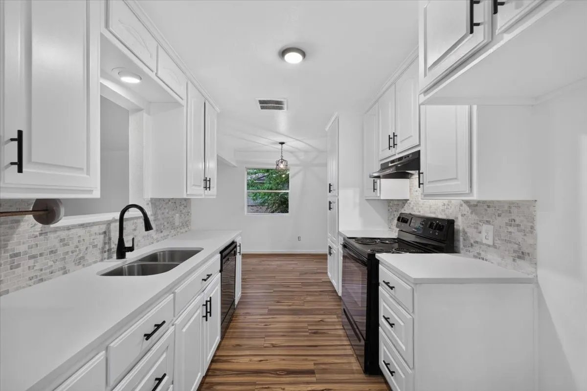 Galley kitchen featuring white cabinetry, matte black hardware, dual basin sink with gooseneck faucet, light-toned backsplash, and dark wood-finish flooring