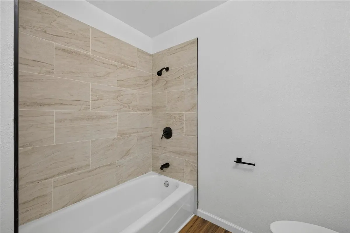 Bathtub with matte black fixtures, light-toned tile surround, and wood-finish flooring