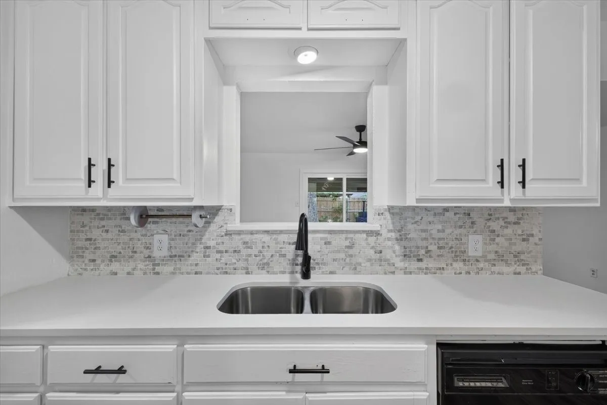 White cabinetry with recessed panel doors, mosaic tile backsplash, double basin stainless steel sink, matte black faucet, and white countertops