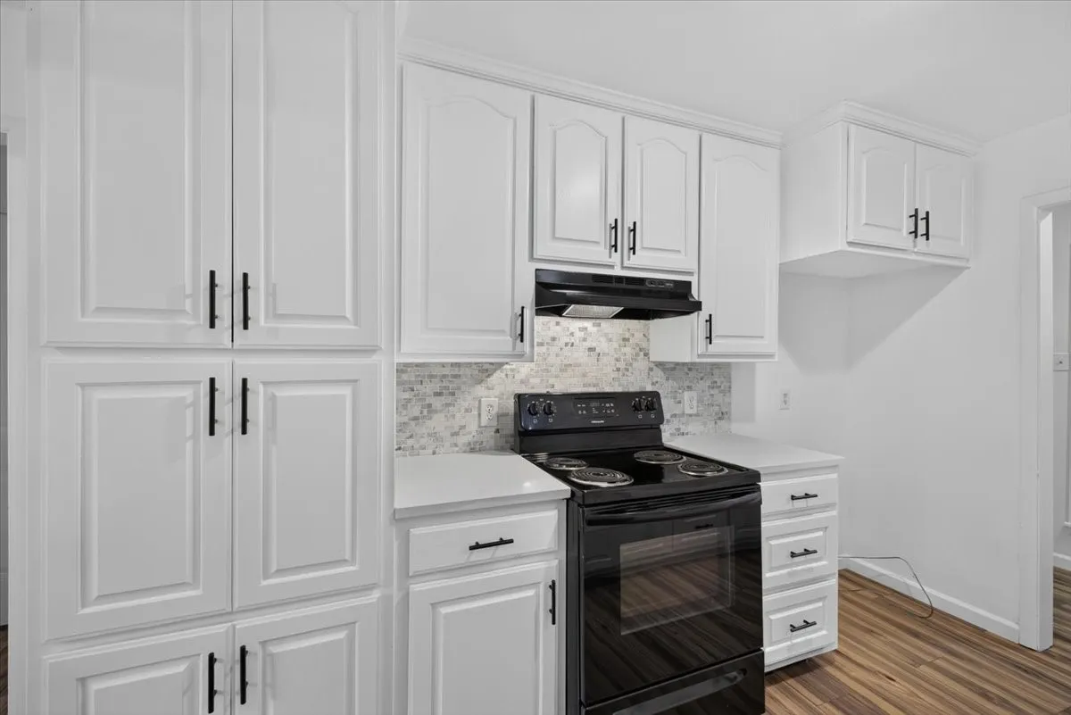 White cabinetry with matte black hardware, mosaic tile backsplash, black electric range with coordinating range hood, and wood-finish flooring
