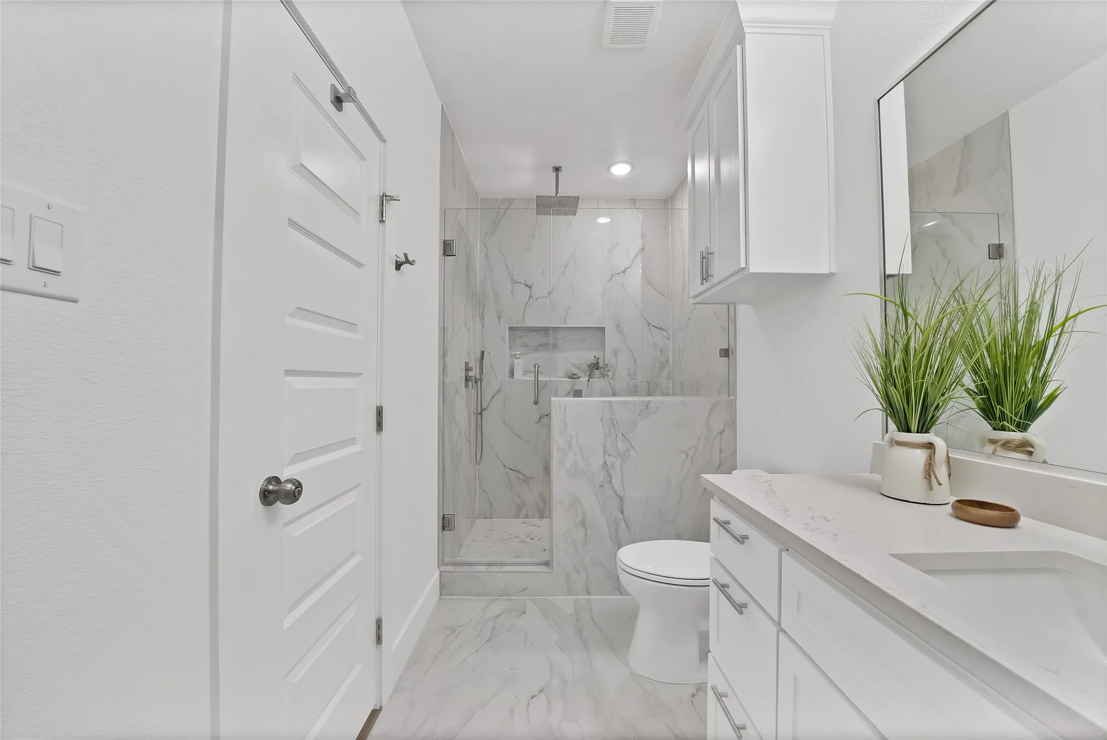 Spacious bathroom featuring a single vanity with an integrated sink, white cabinetry, a large mirror, and a walk-in shower with stone tile surround and glass enclosure