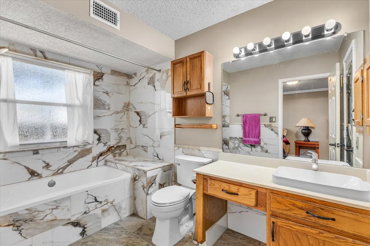 Bathroom featuring a bathtub, toilet, wood-finish vanity with an integrated vessel sink, a large wall-mounted mirror, and tile flooring